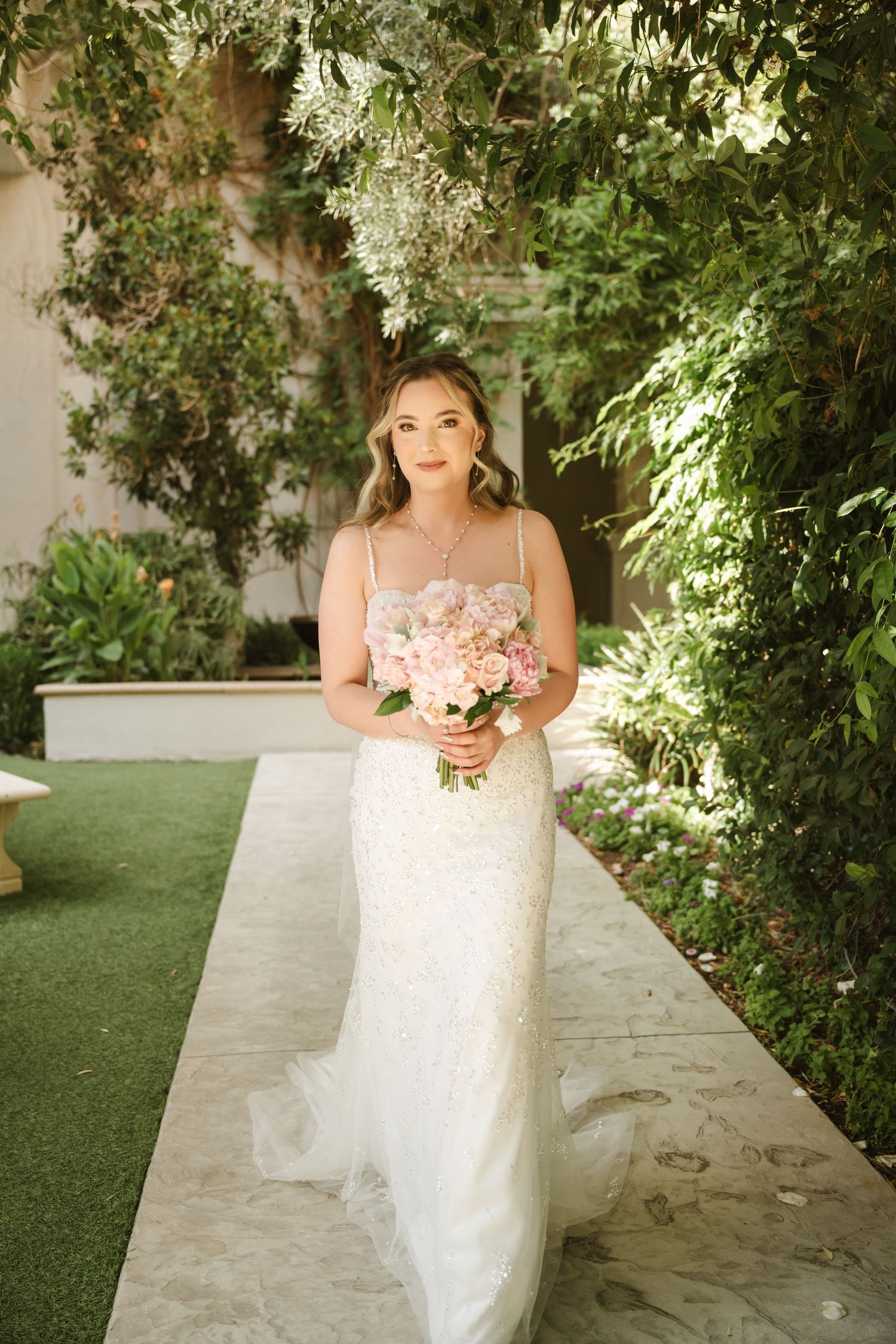 A bride holds a pink bouquet while standing in a lush garden setting.