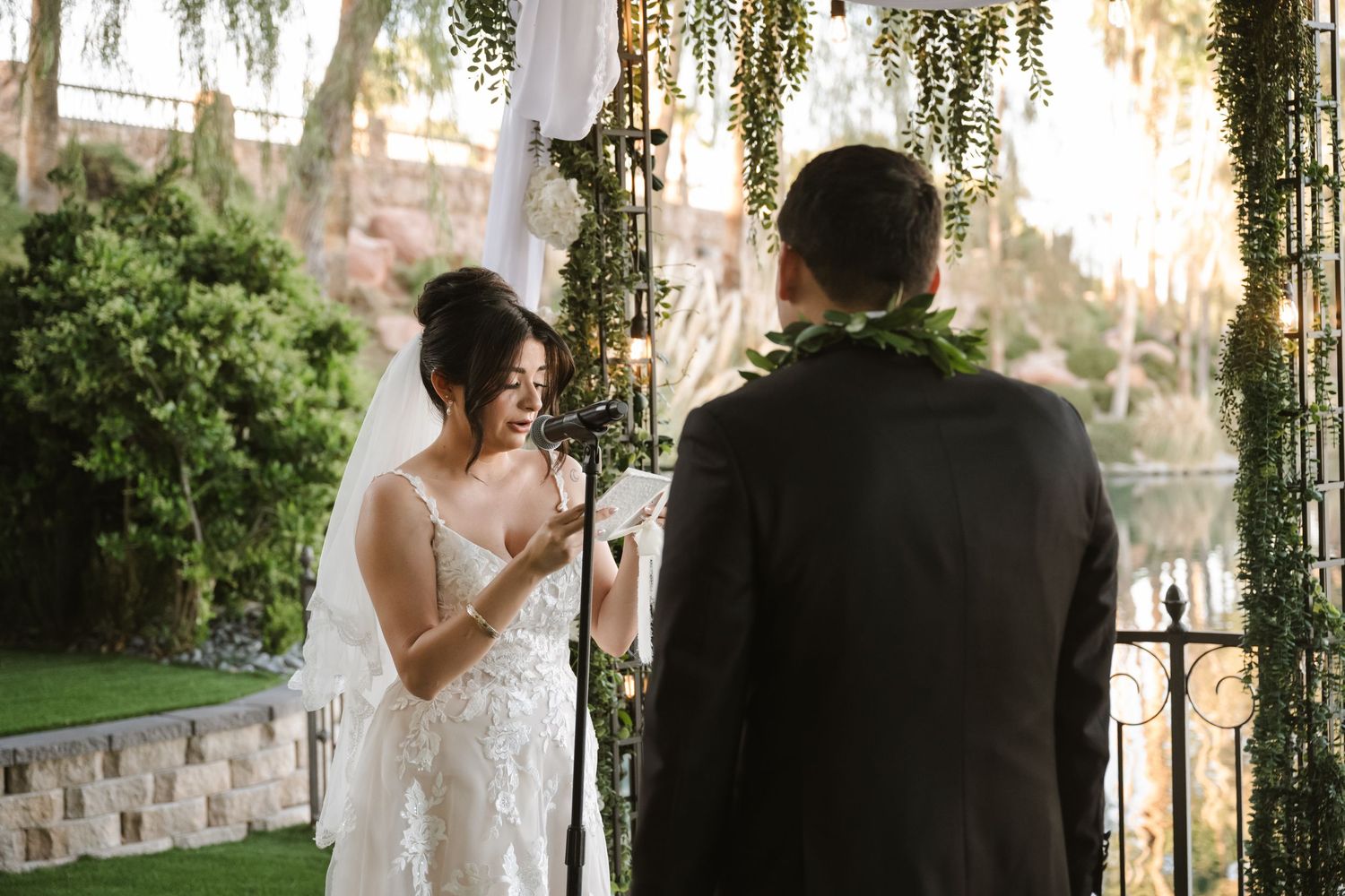 Bride smiles joyfully at groom during outdoor wedding ceremony under a lush greenery-draped arch.