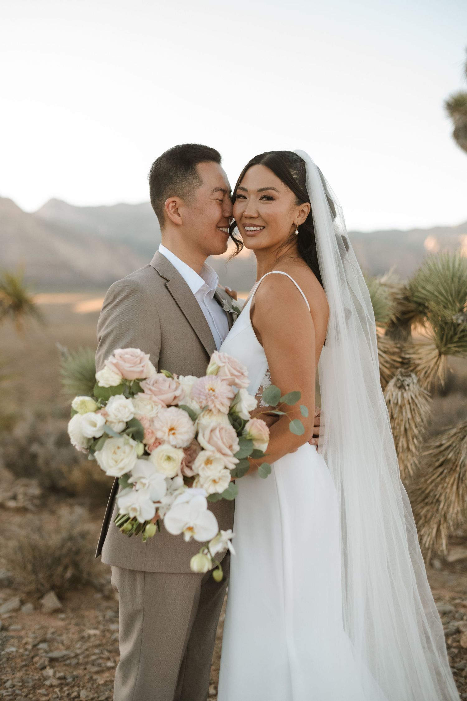 Bride and groom pose in desert landscape with Joshua trees, she holds blush bouquet, he wears tan suit.