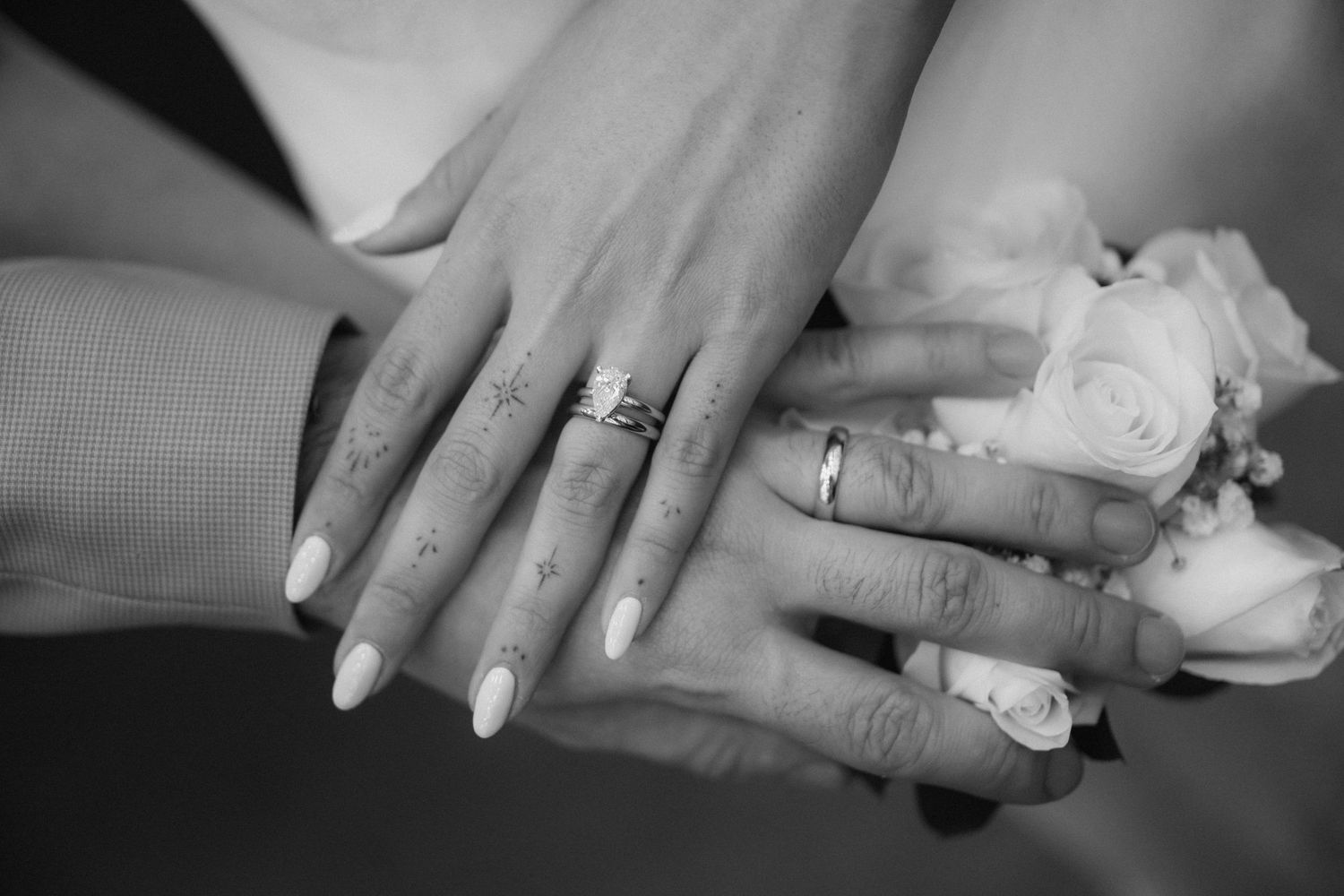 Black and white photo of two brides showing off wedding rings and manicured nails over a bouquet of roses.