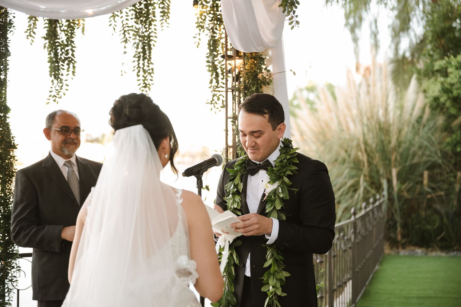 Groom in tuxedo with floral lei reads wedding vows to bride in white gown at outdoor ceremony.