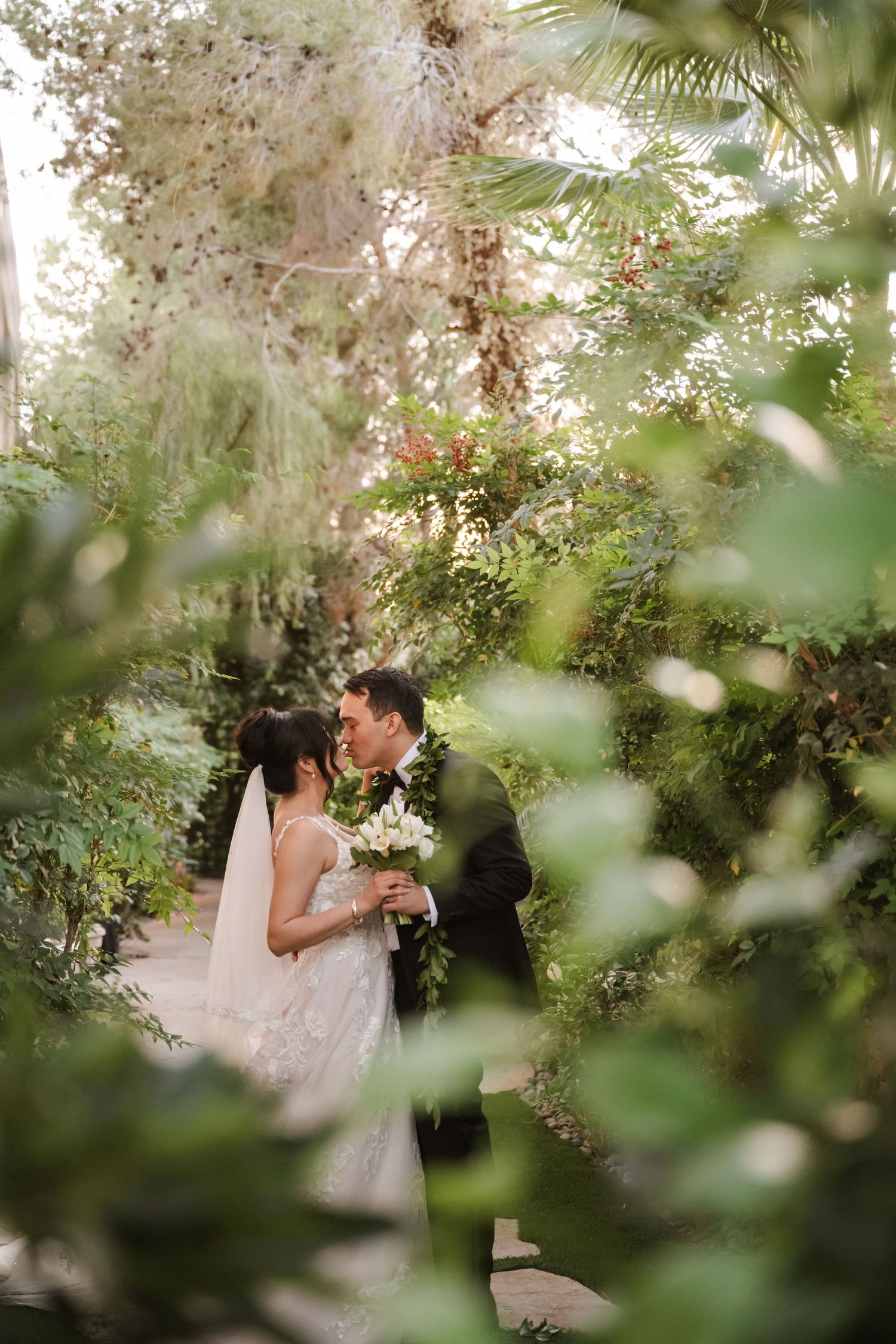 A bride and groom share a romantic kiss surrounded by lush green garden foliage on their wedding day.