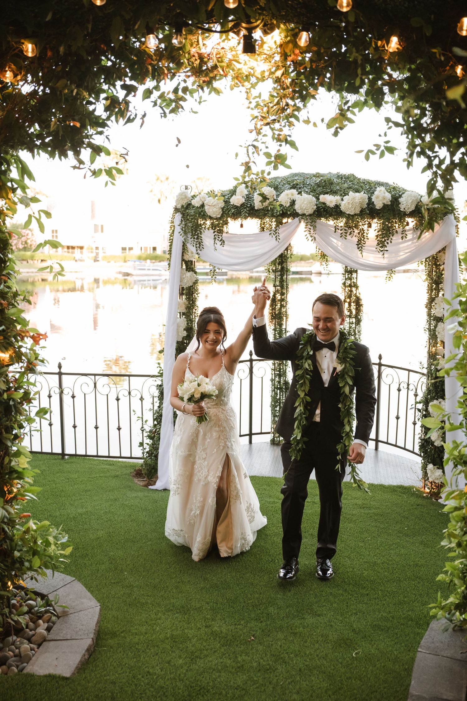 Happy bride and groom raise hands in celebration under greenery arch at lakeside wedding.