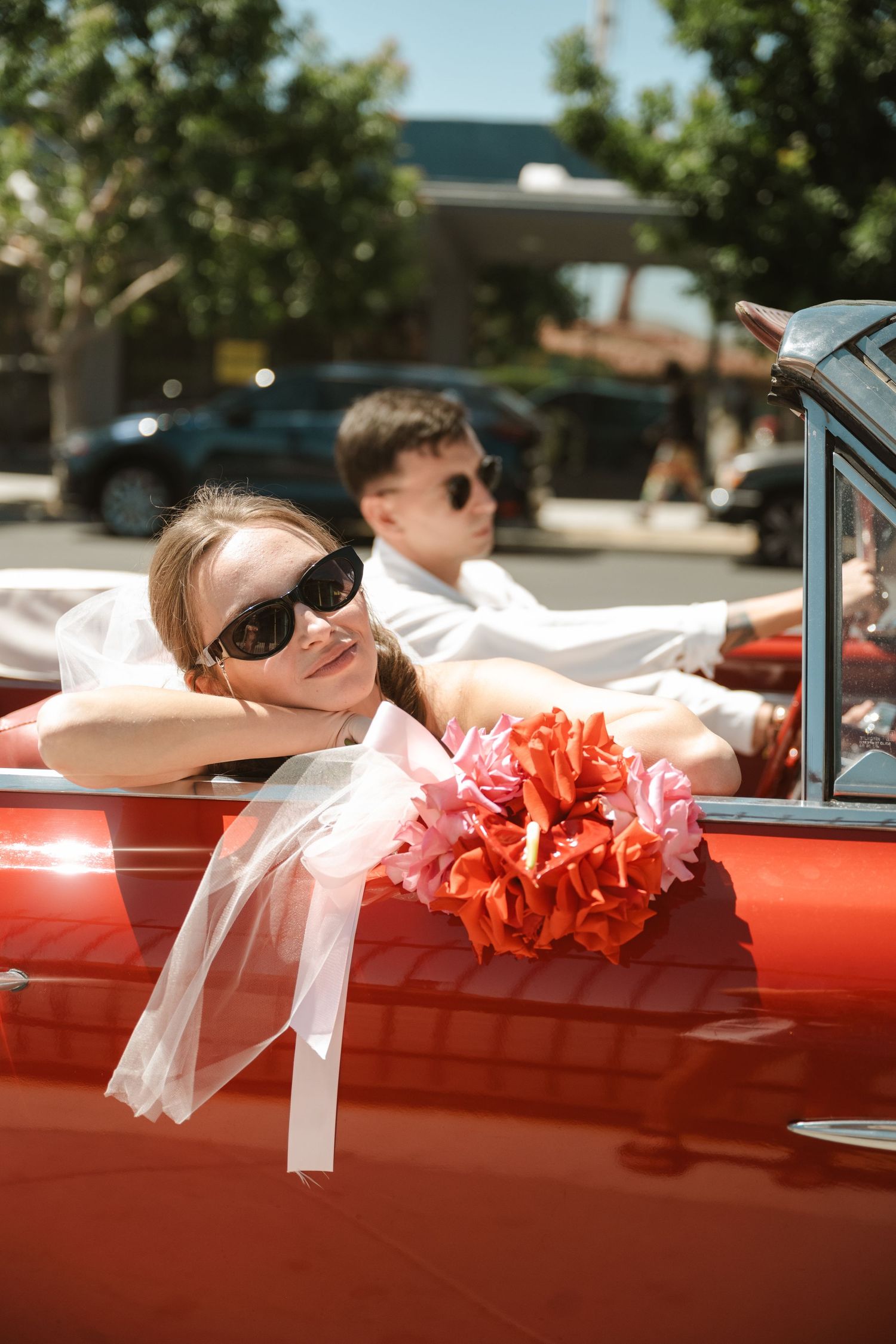 Newlywed couple in sunglasses riding in a classic red convertible car with colorful floral bouquet on a sunny day.