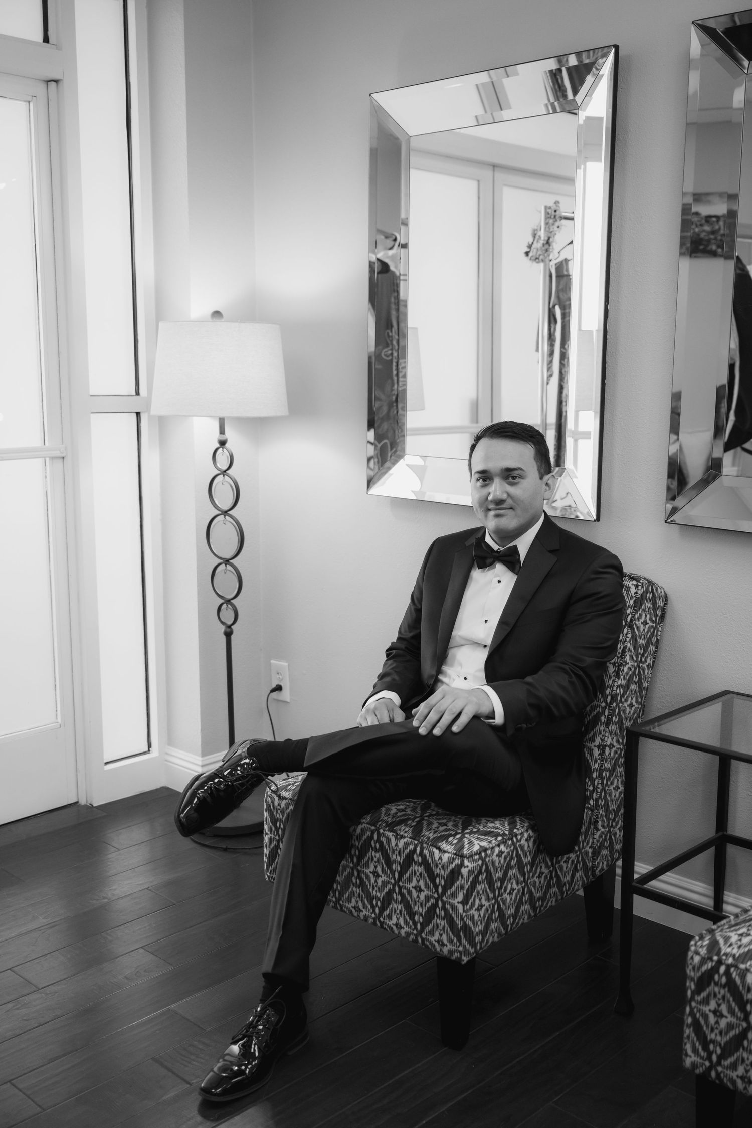 A man in a tuxedo sits in a patterned chair in a elegant room with a mirror and floor lamp.