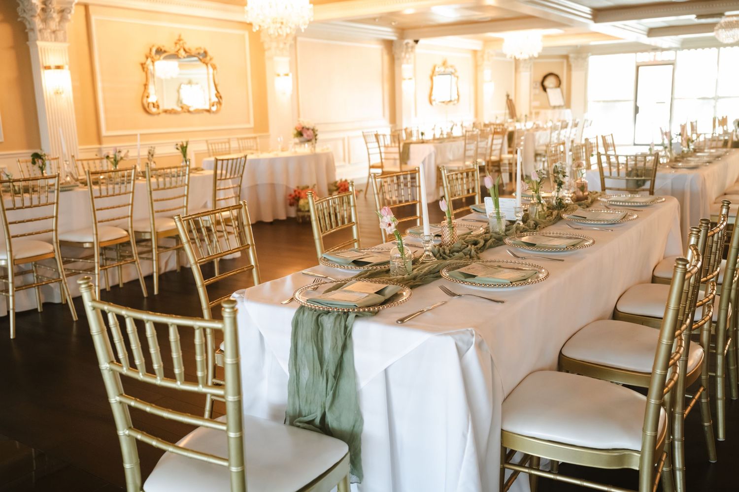Elegant wedding reception hall with gold chiavari chairs, long white tables, sage green runners, and chandelier lighting.