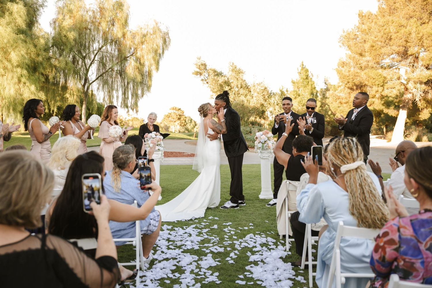 Outdoor wedding ceremony with couple kissing at altar, guests applauding, white flower petal aisle, lush green trees backdrop.