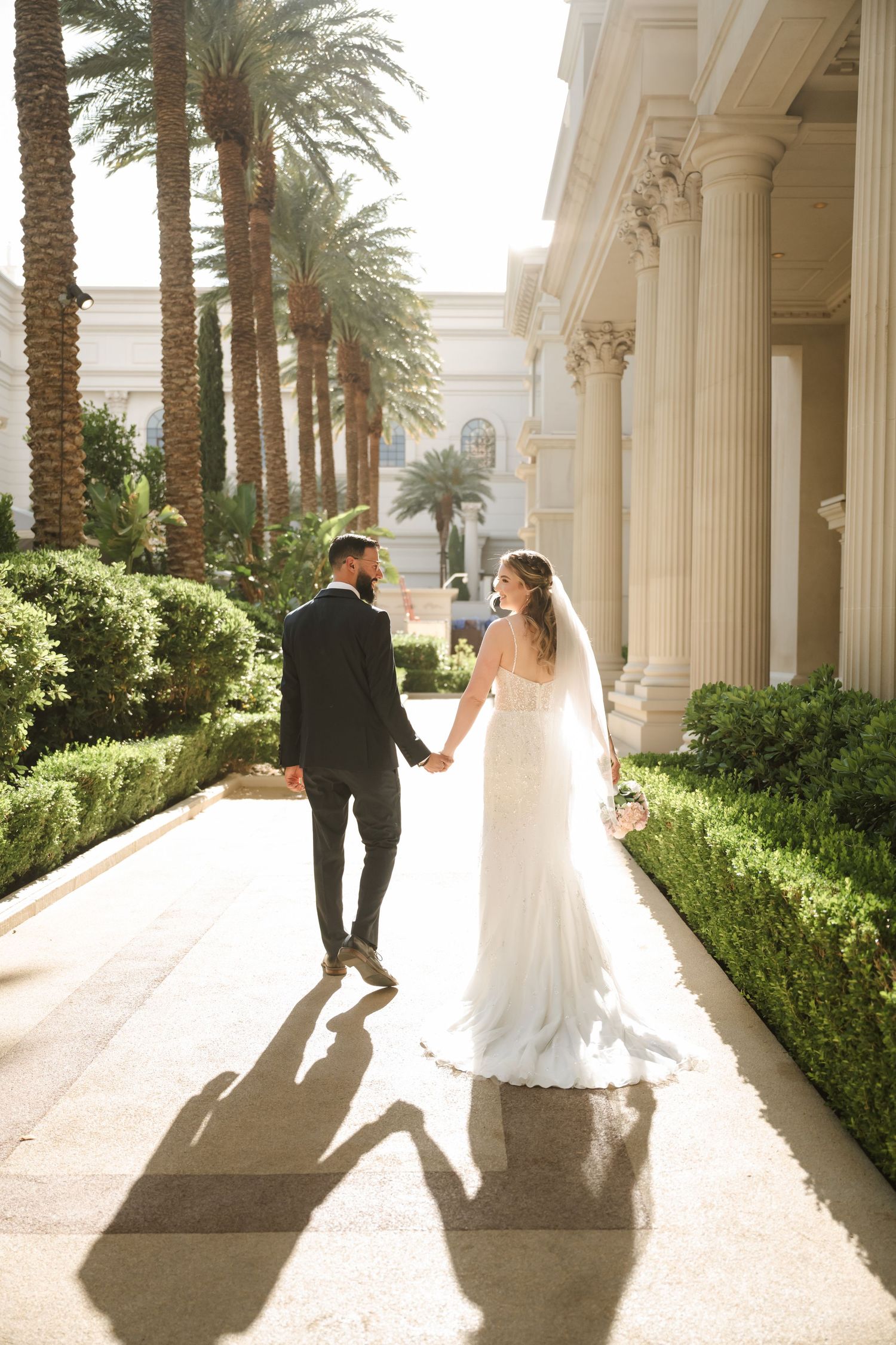Bride and groom walk hand-in-hand along a sunlit path with palm trees and white columns at an elegant venue.