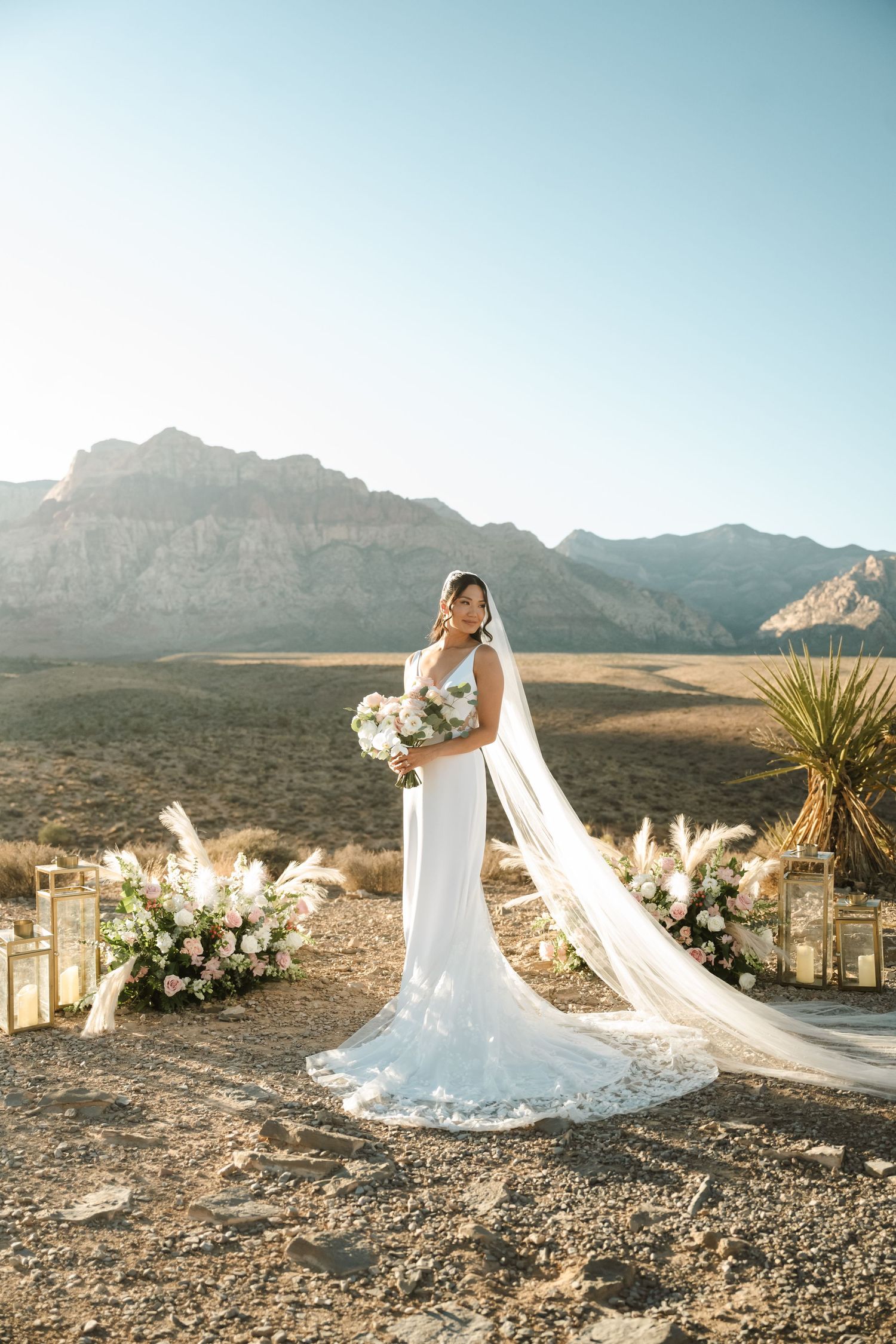 Bride in white gown with long veil holds bouquet at desert ceremony with floral arrangements and Red Rock Canyon backdrop.