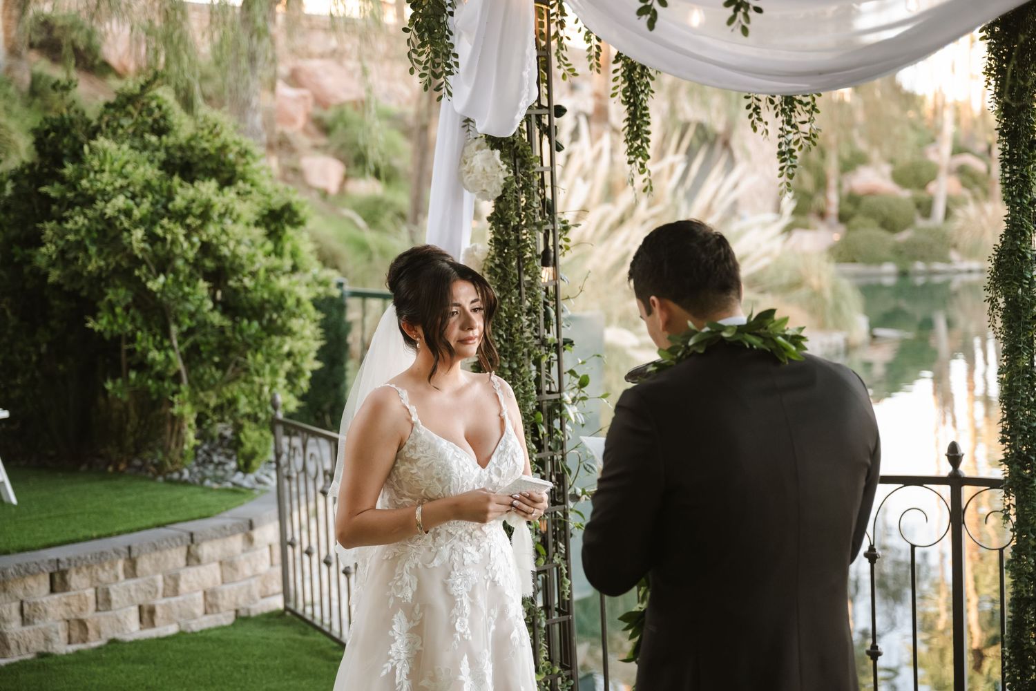 Bride smiling at groom during outdoor wedding ceremony under floral arch with greenery and water view.