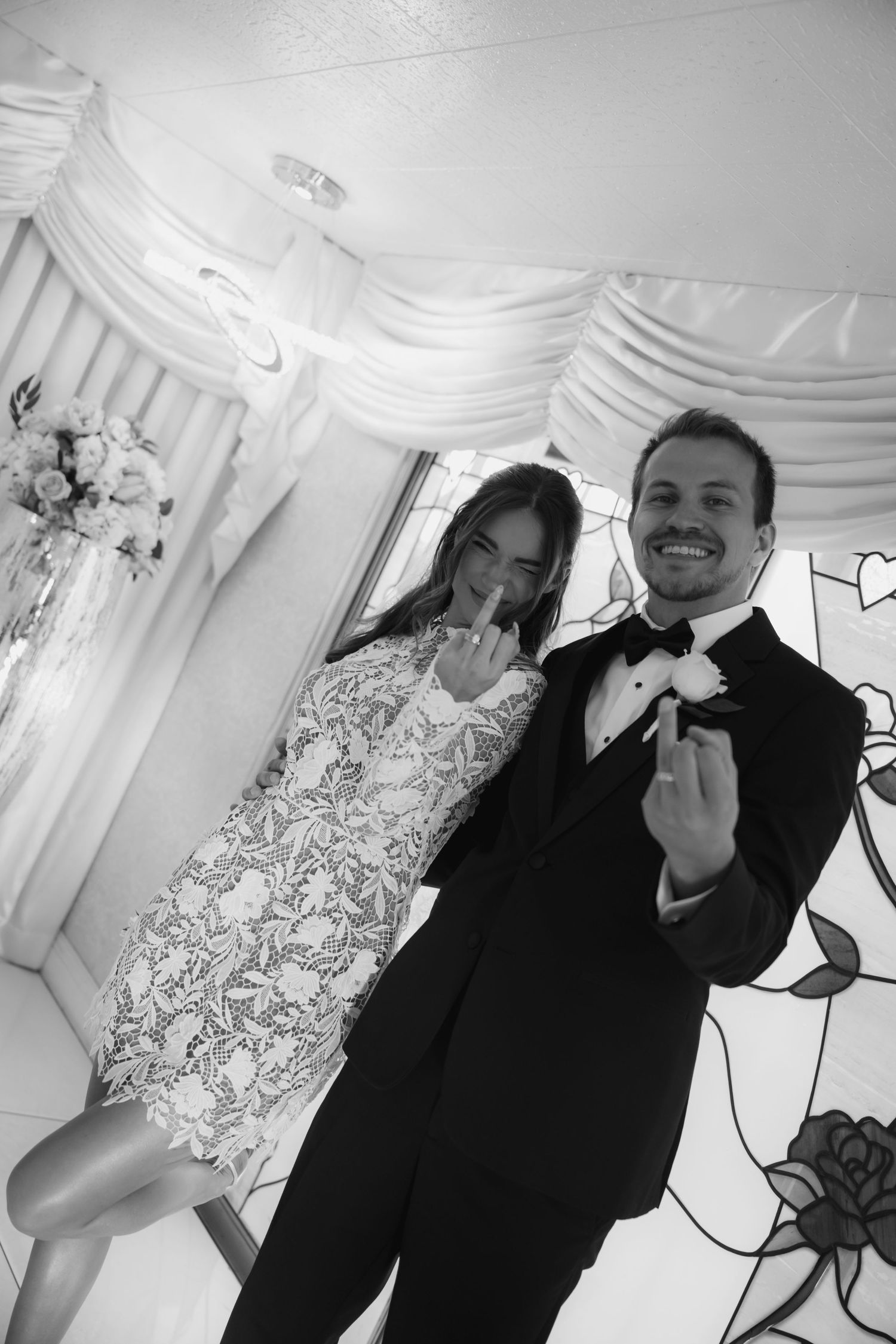 Black and white photo of smiling newlyweds in wedding chapel, both showing off wedding rings, groom in tuxedo.