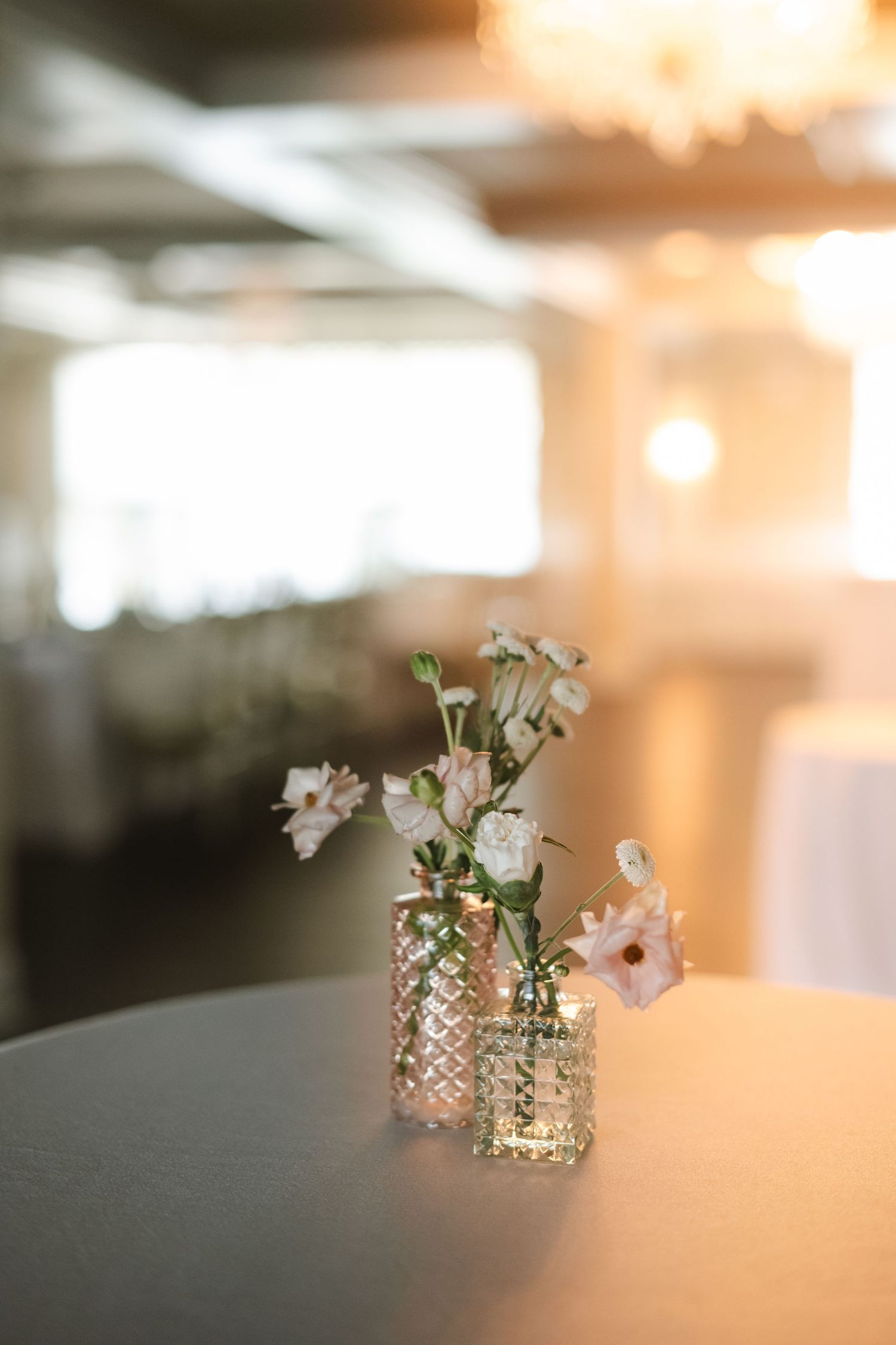 Two small crystal bud vases with delicate white and pink flowers on a table at an elegant wedding reception.