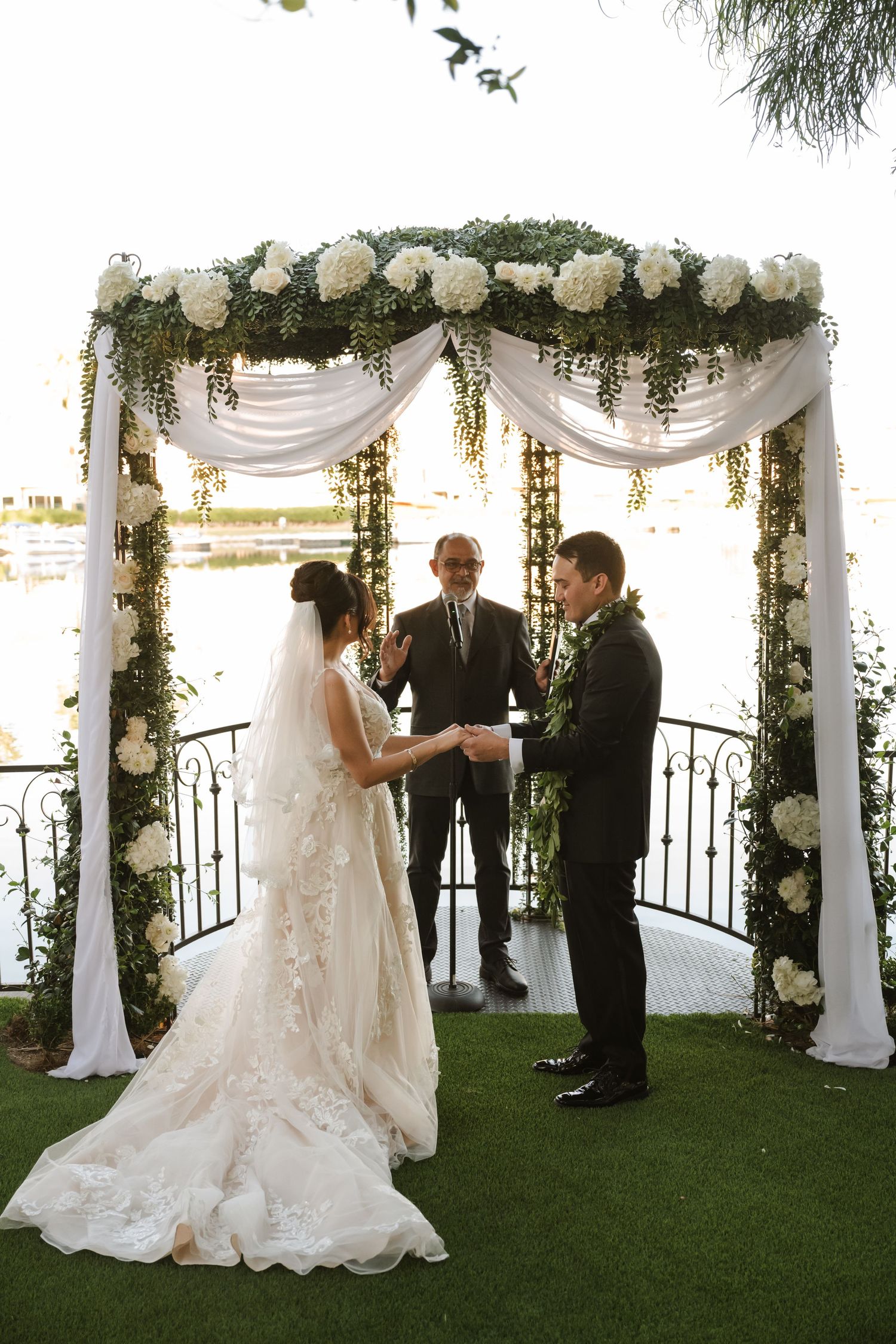 Bride and groom exchange vows under floral arch with white roses and greenery at outdoor waterfront wedding ceremony.