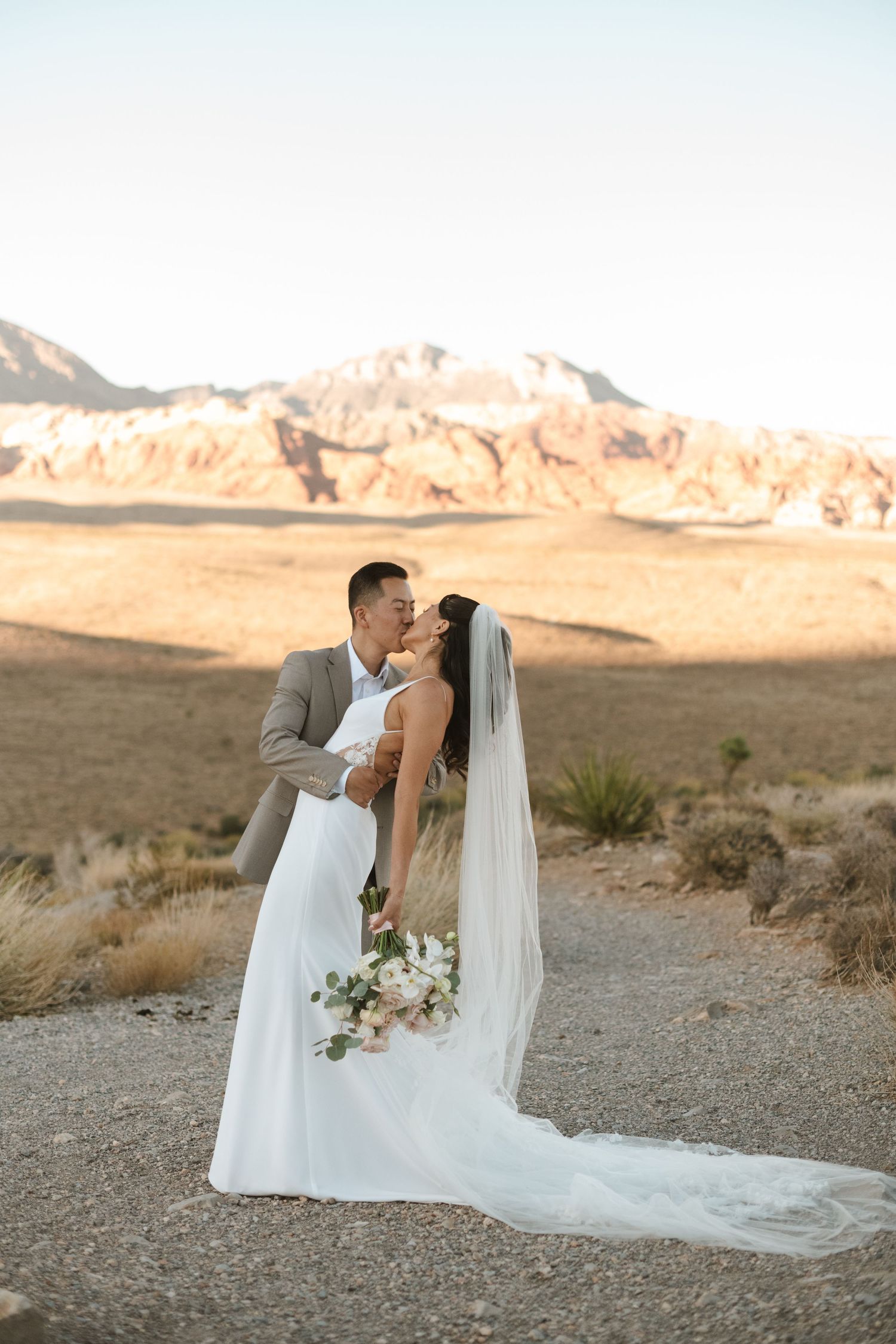 Bride and groom kissing at Red Rock Canyon desert landscape, bride in white gown with veil holding floral bouquet.