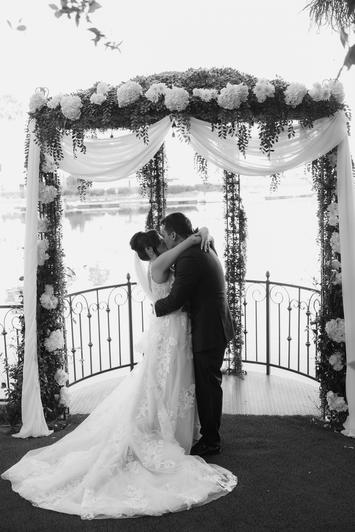 Black and white wedding photo of couple kissing under elegant floral arch near waterfront.