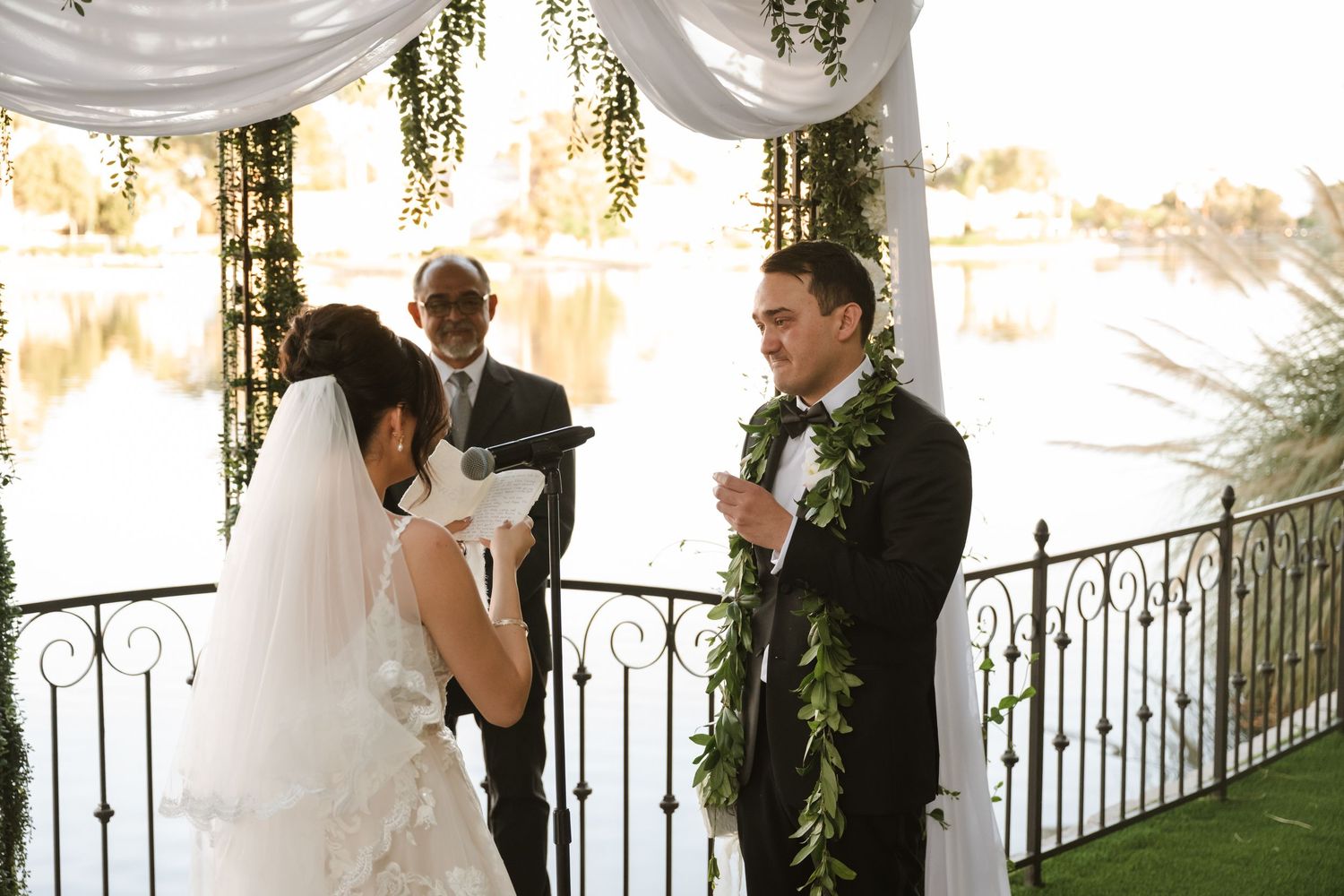 Bride and groom exchange vows at outdoor waterfront wedding ceremony under floral arch with officiant.