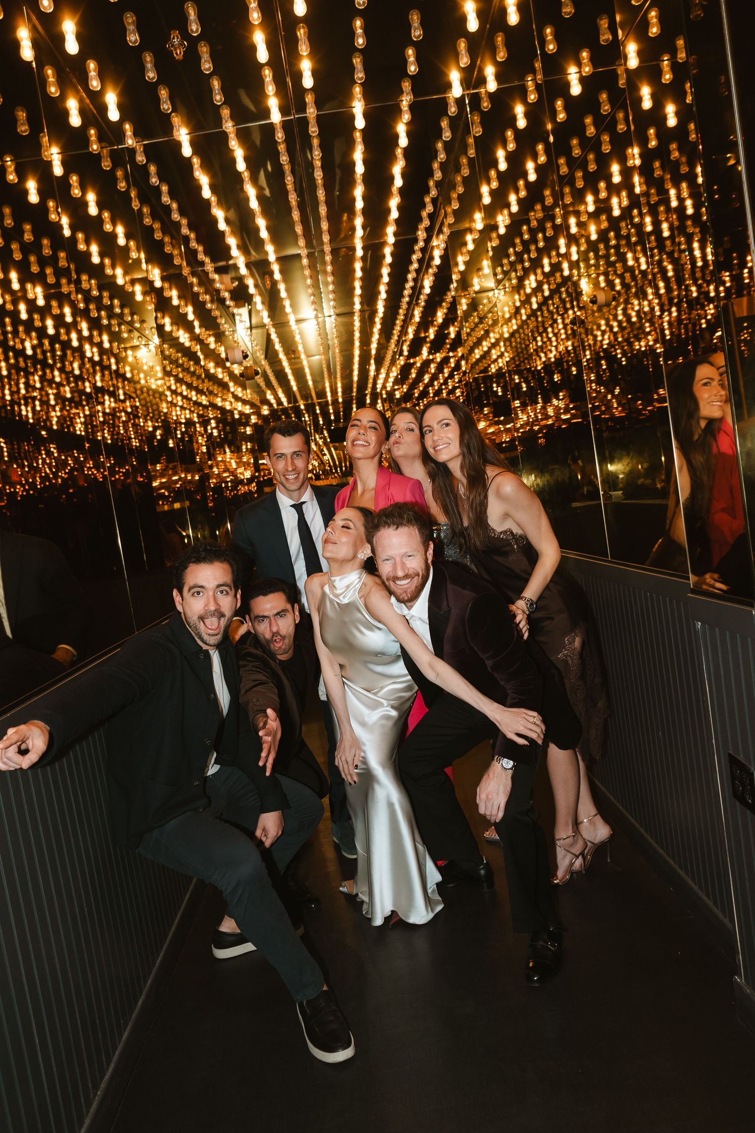 Group of elegantly dressed friends posing together under warm string lights at a glamorous evening event.