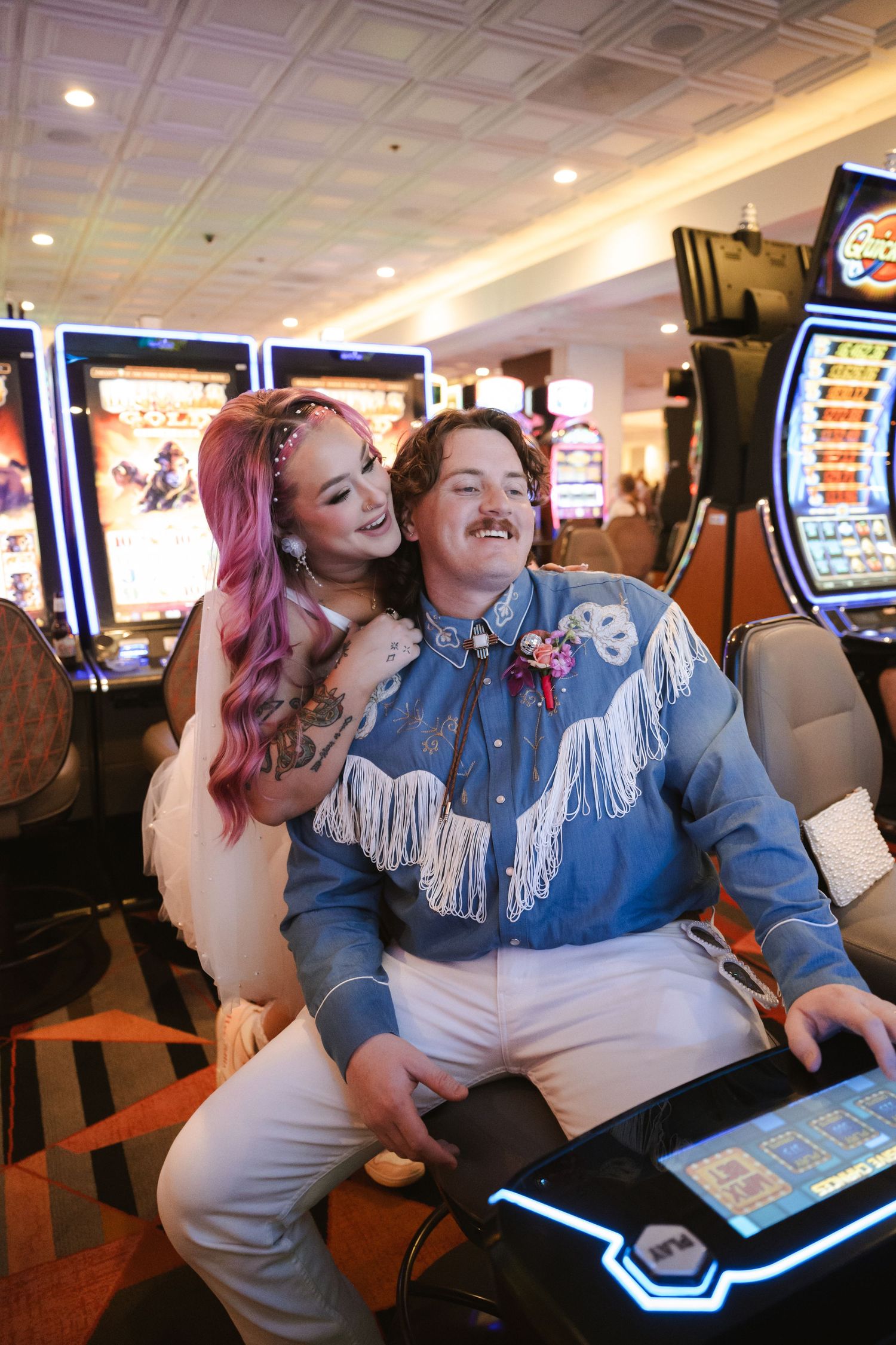 Couple laughing together at a casino slot machine, woman with pink hair hugging man in blue fringe western shirt.
