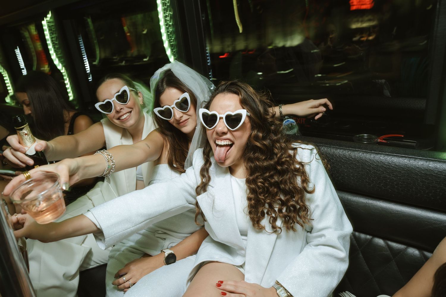 Three women in white outfits and heart-shaped sunglasses celebrate a bachelorette party inside a party bus.