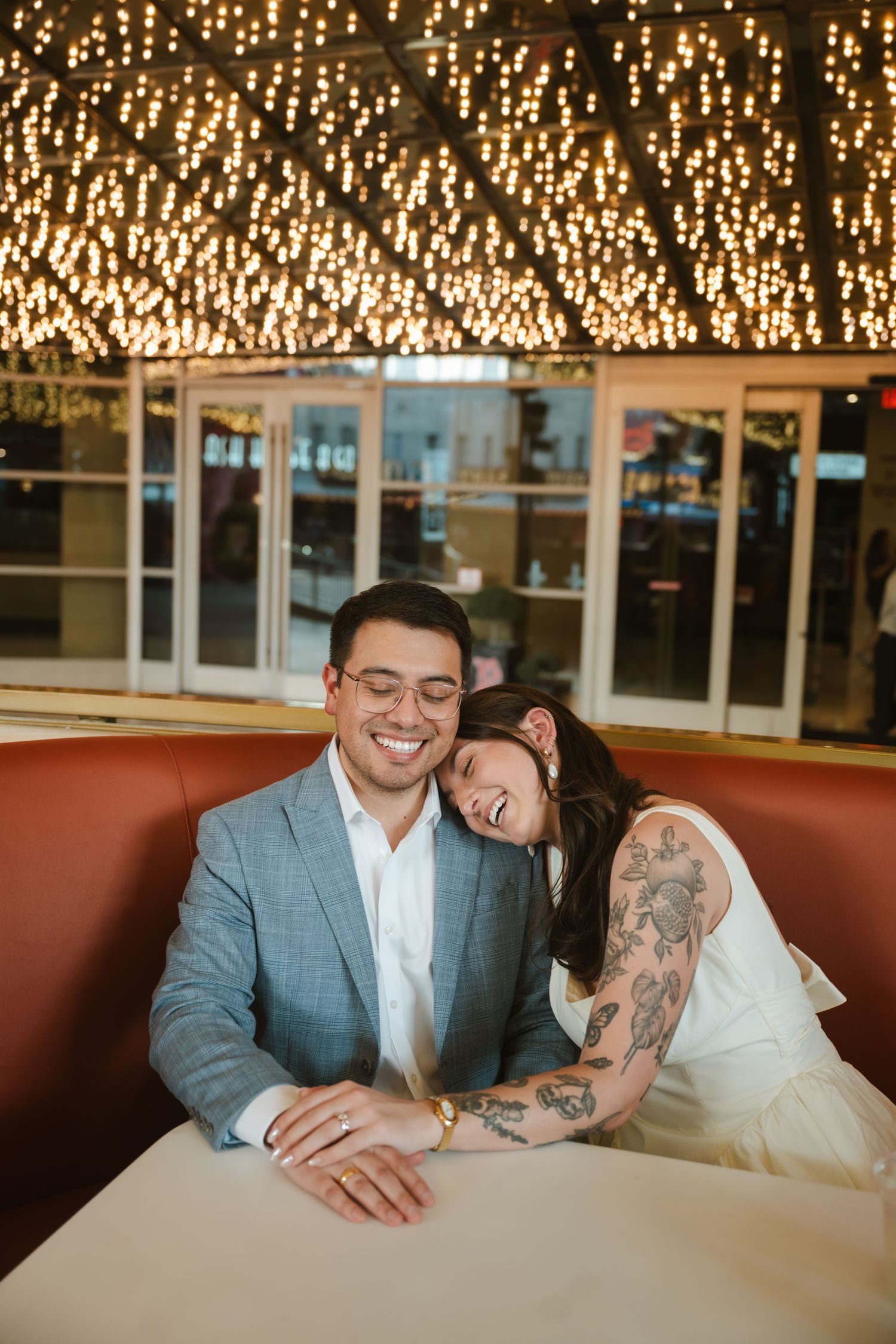 Couple sharing a joyful moment in a restaurant booth, woman with tattoos leaning on man in gray suit under warm lights.