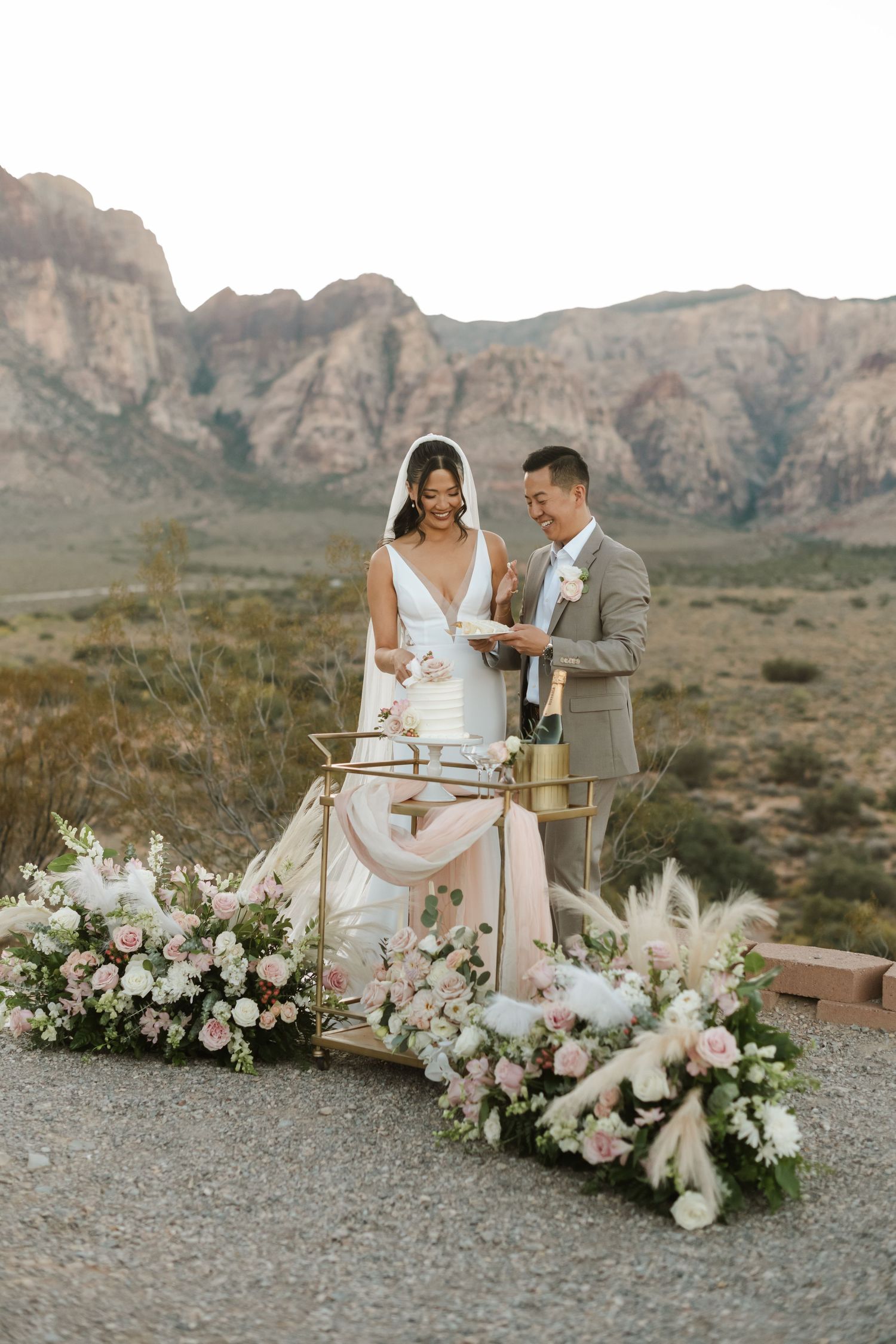 Couple cuts wedding cake outdoors at Red Rock Canyon, surrounded by pink floral arrangements and desert mountains.