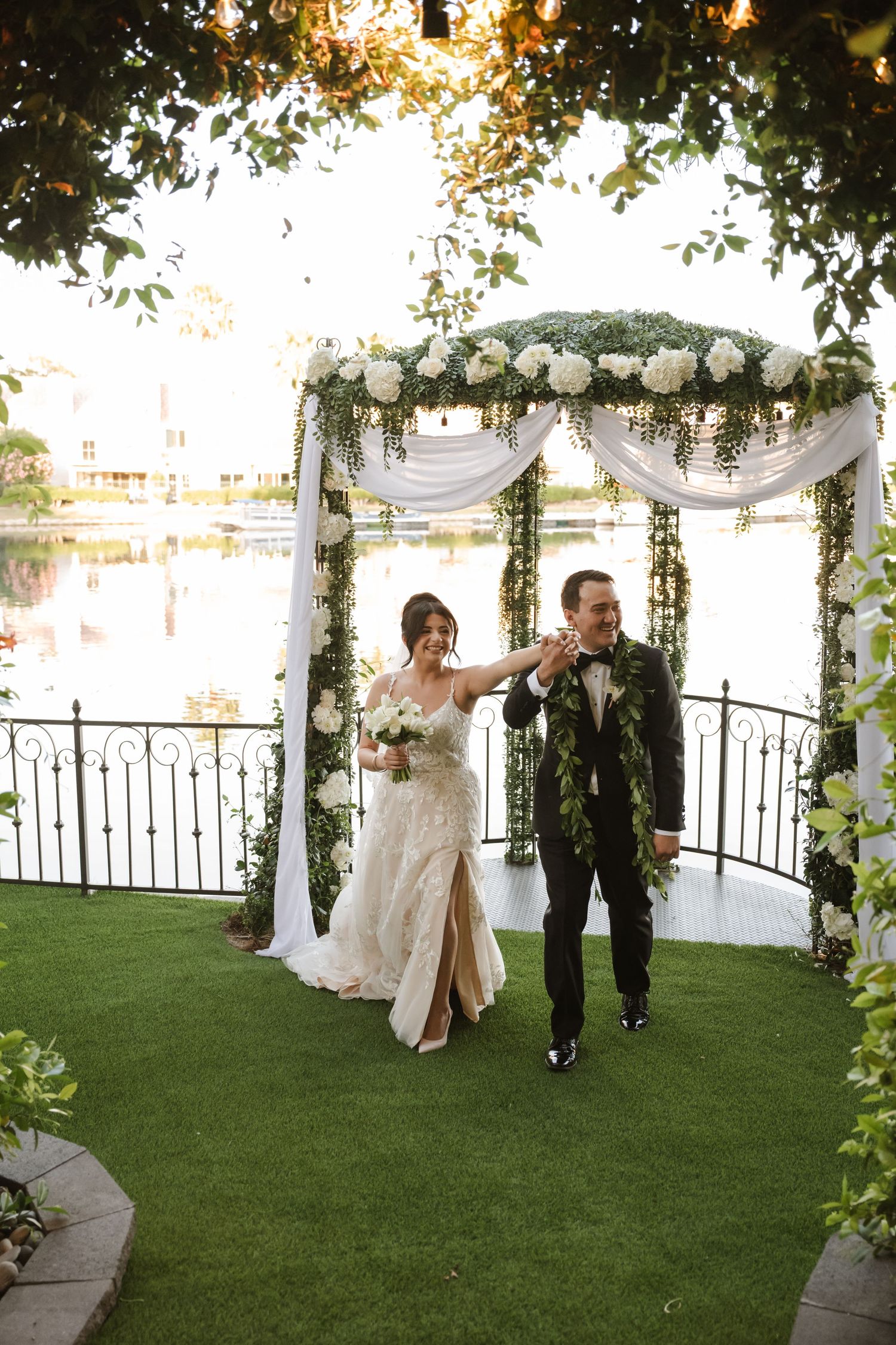 Joyful newlyweds walk hand in hand under floral arch after outdoor waterfront wedding ceremony.