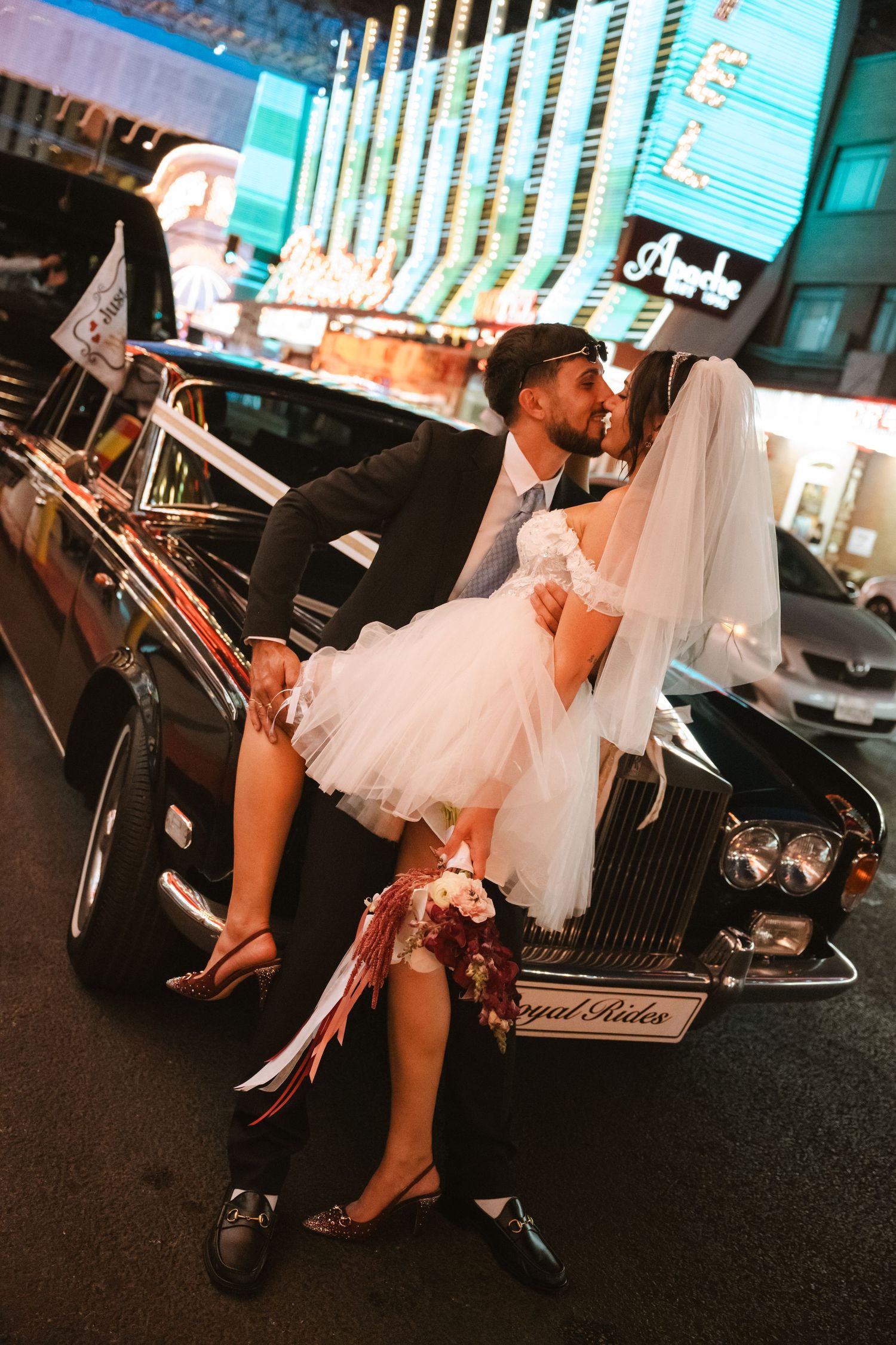 Newlywed couple kissing beside a classic black Rolls-Royce on a vibrant Las Vegas strip at night.
