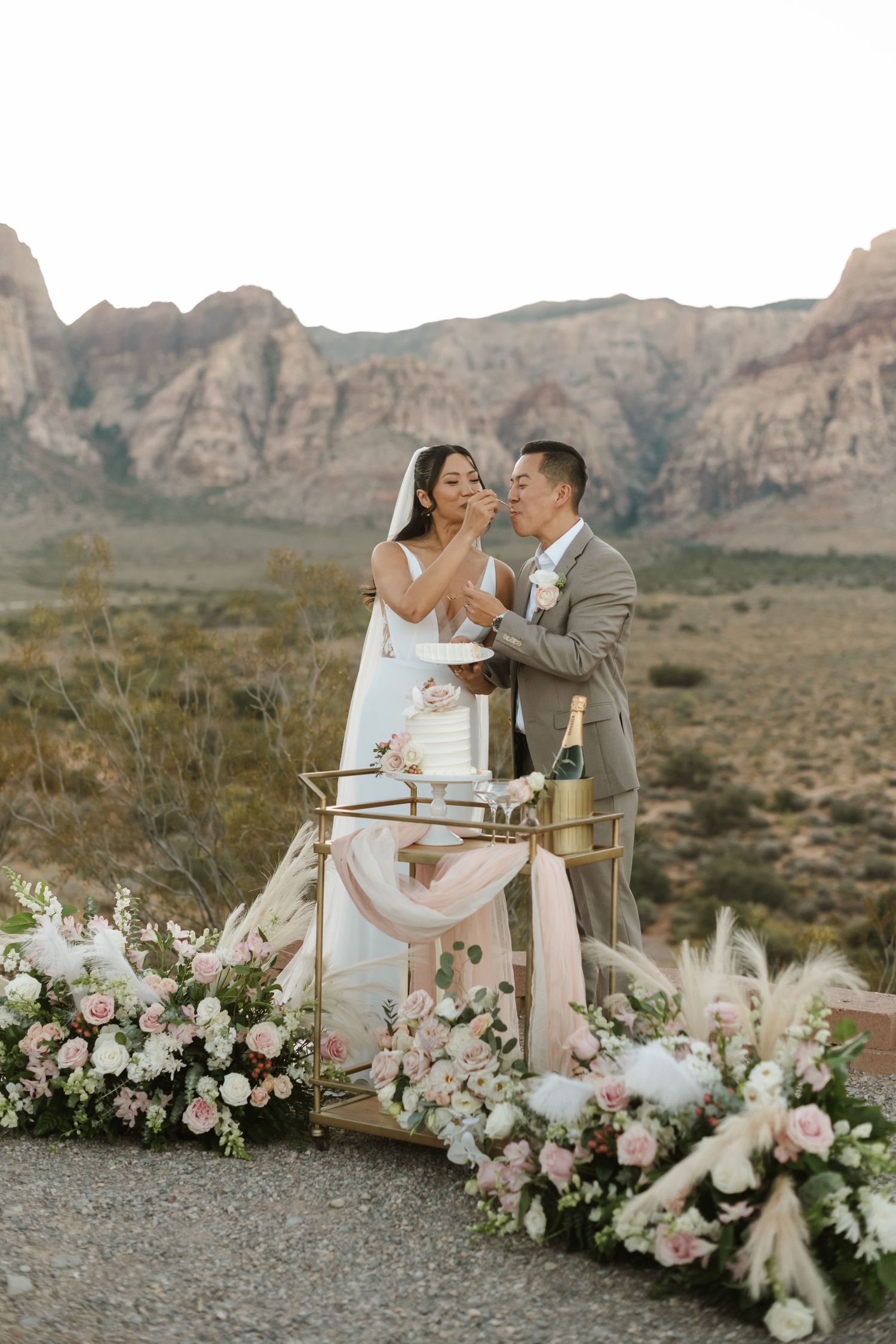 Couple cutting wedding cake outdoors at Red Rock Canyon, surrounded by pink floral arrangements and desert mountains.