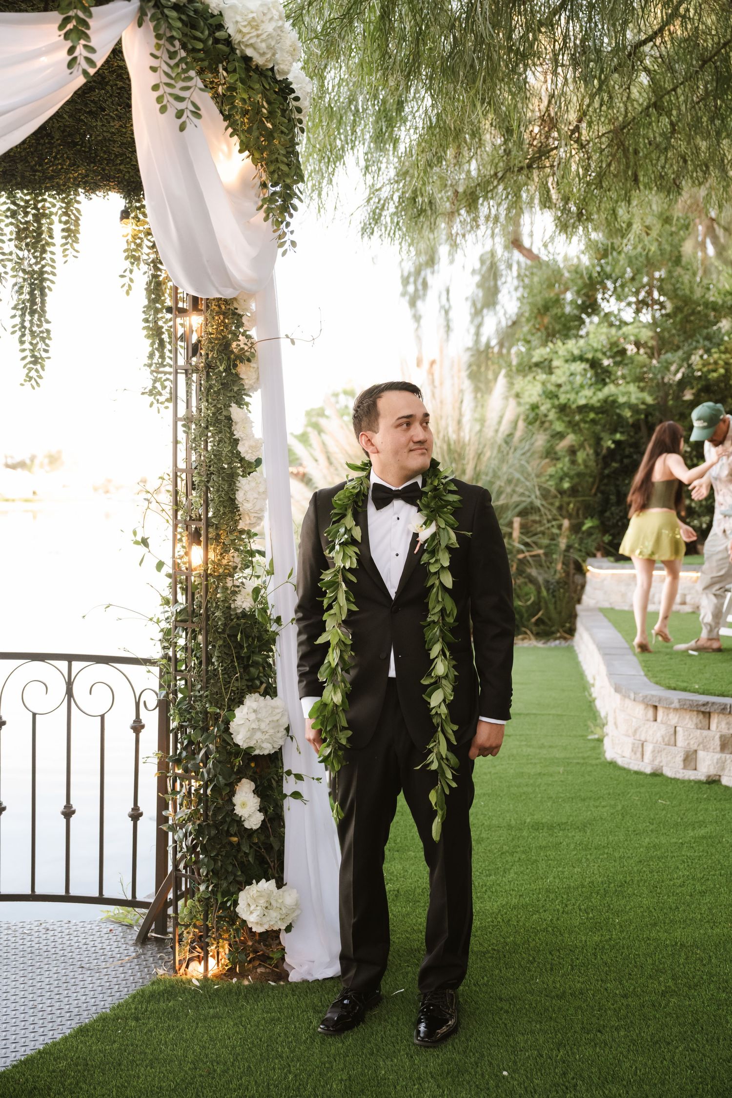 Groom in black tuxedo with green leaf lei stands at outdoor wedding altar decorated with white flowers.