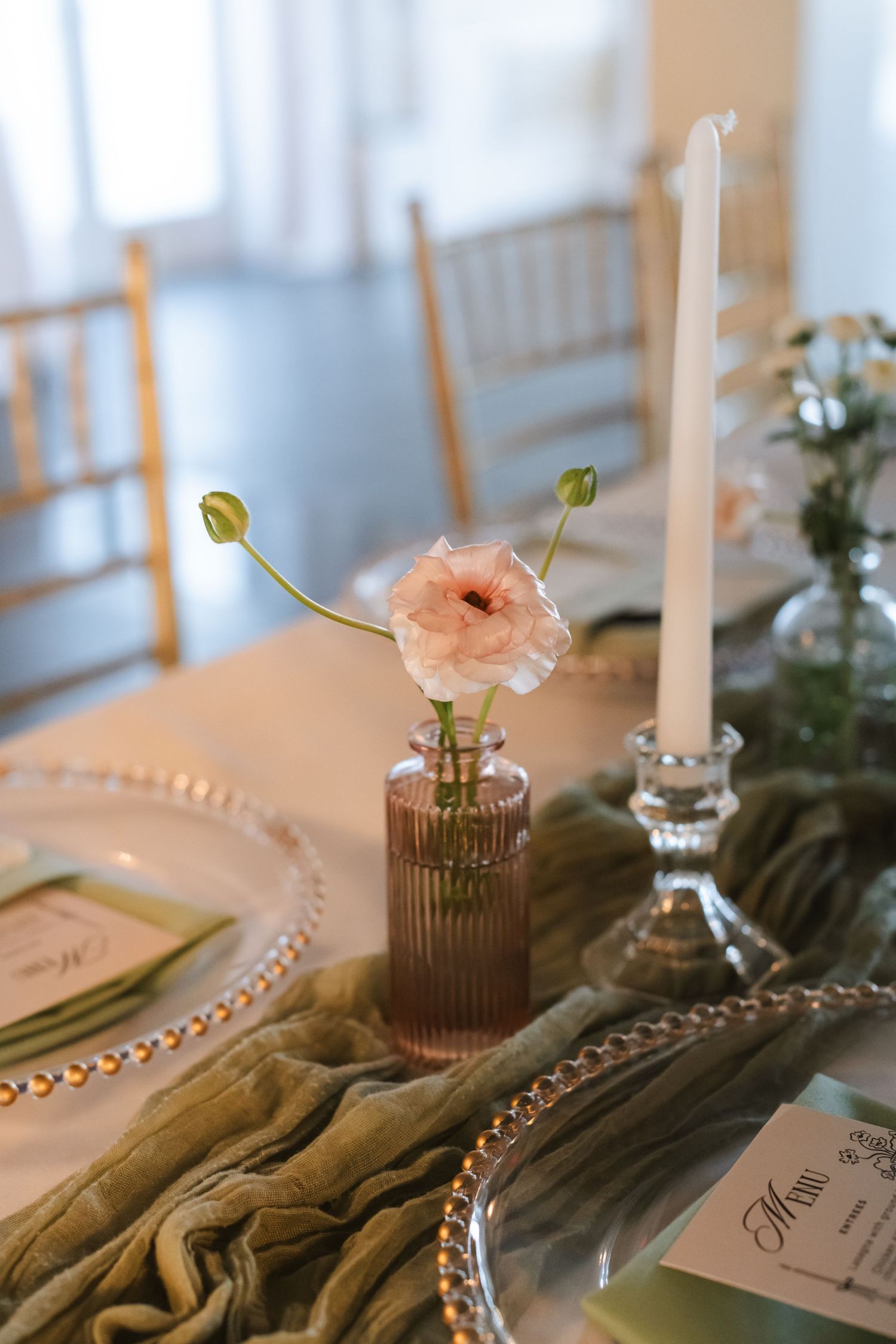 Elegant wedding table setting with pink ranunculus in ribbed vase, taper candle, beaded charger plates, and sage green linen runner.