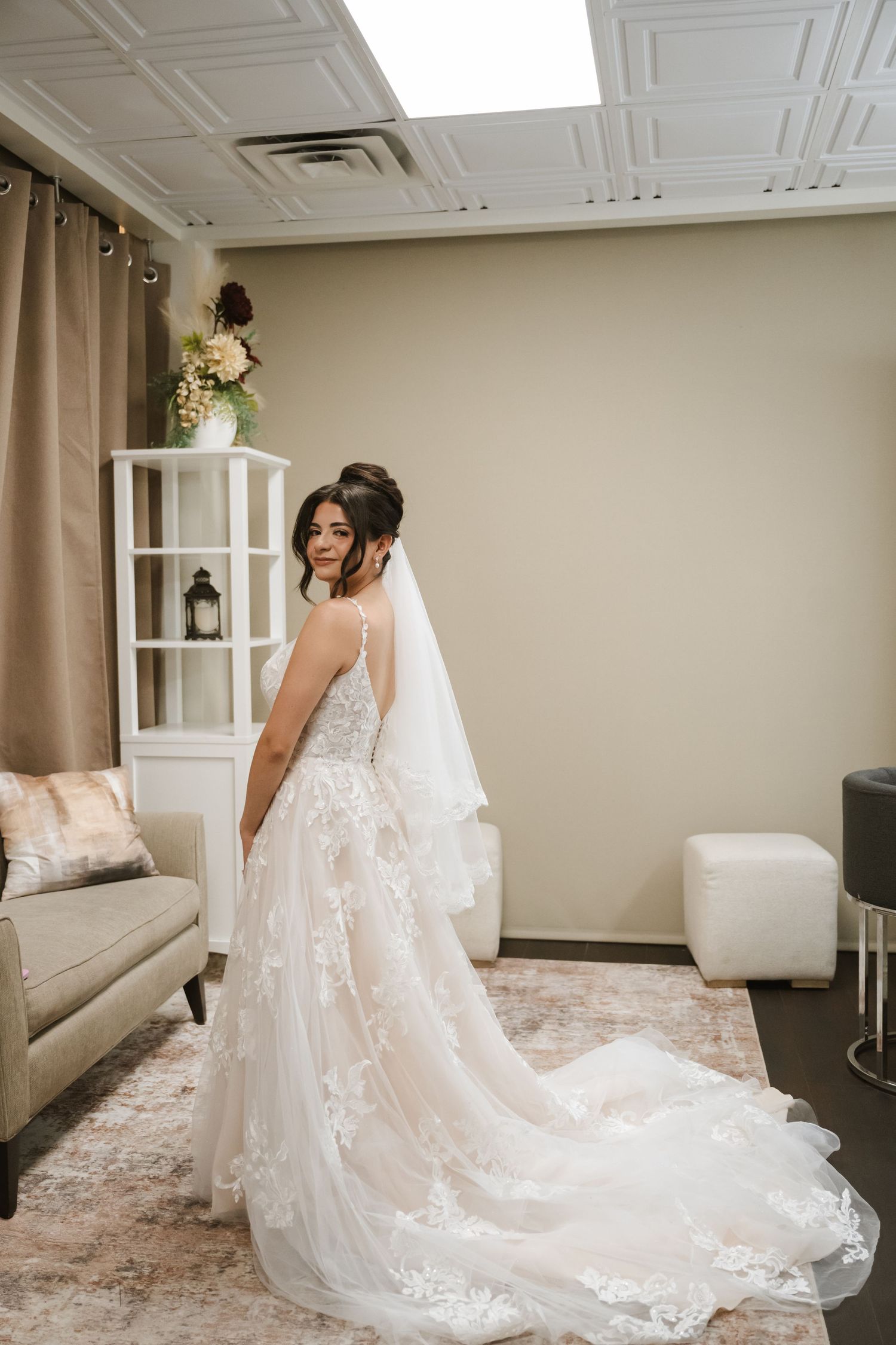 Bride in elegant lace wedding gown with long veil and train poses in bridal boutique fitting room.
