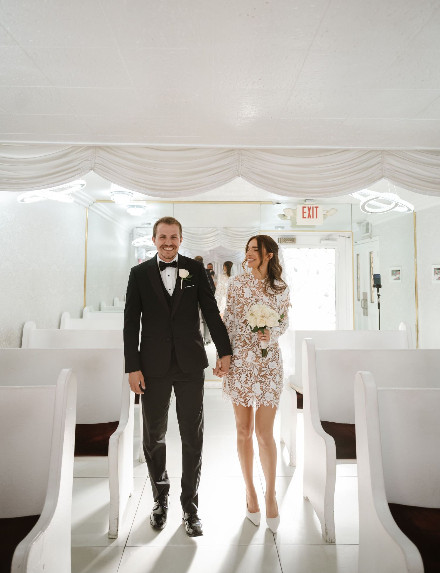 Bride in lace mini dress and groom in black tuxedo walking down aisle of elegant white wedding chapel.