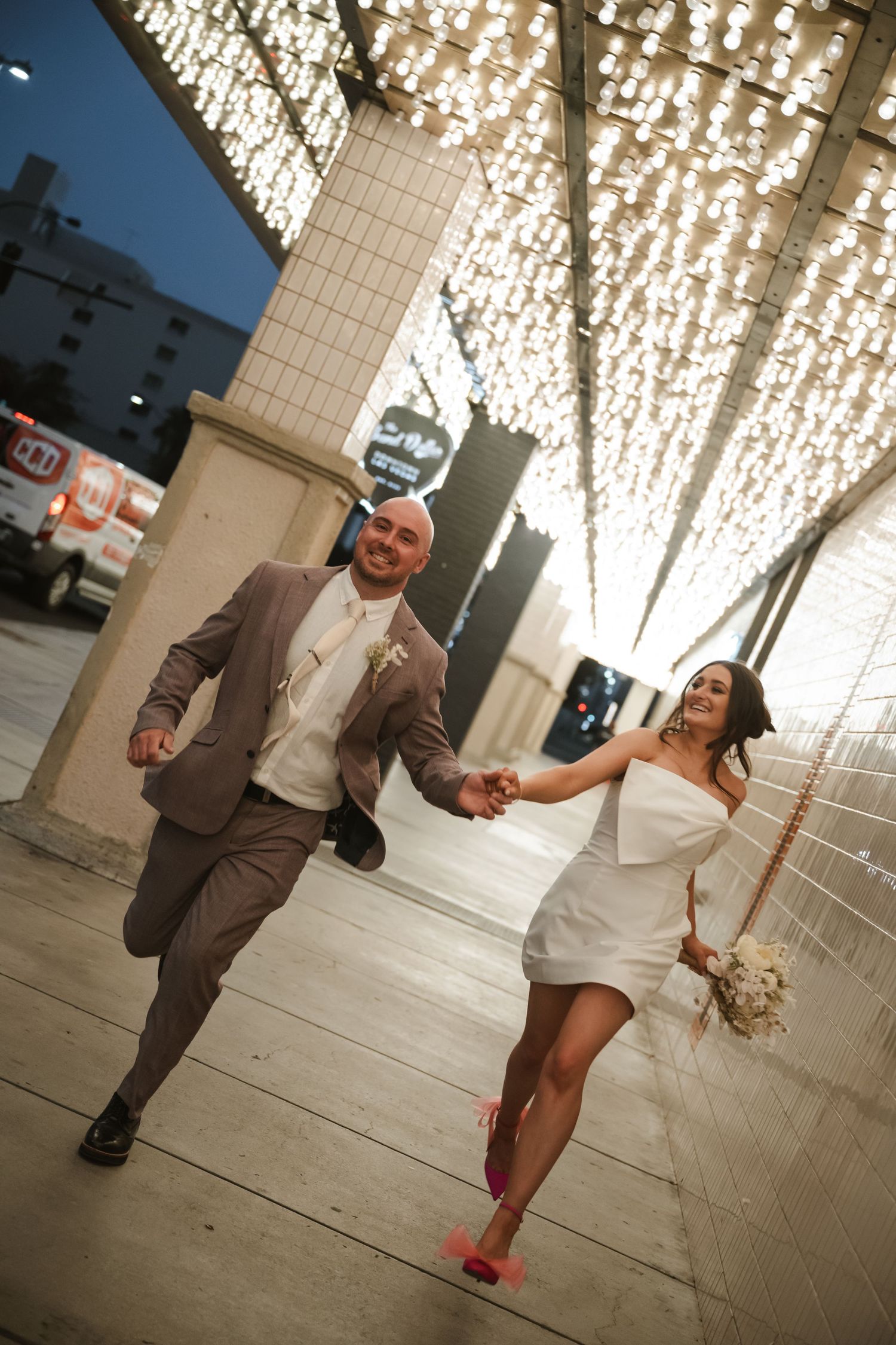 Joyful newlyweds holding hands and running under twinkling fairy lights on a city sidewalk, bride in short white dress.