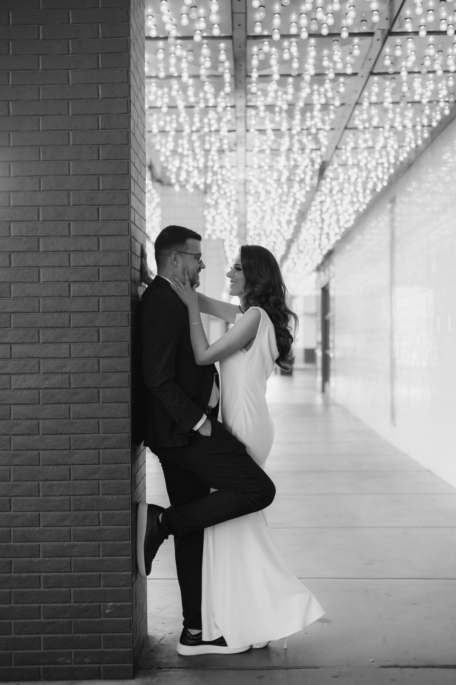 A couple poses creatively against brick walls under sparkling ceiling lights.