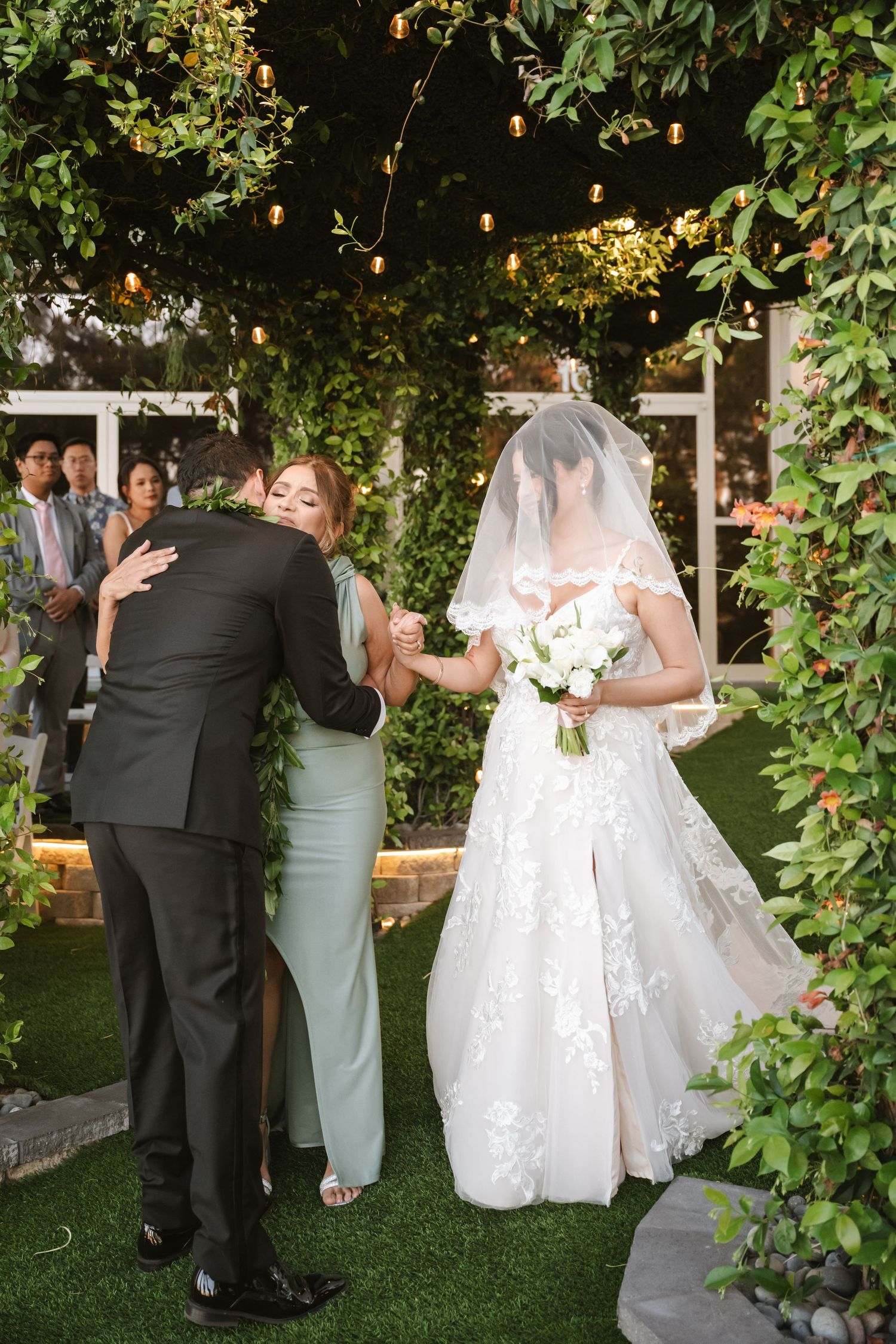 Bride in lace gown with veil walks down outdoor aisle holding white bouquet.