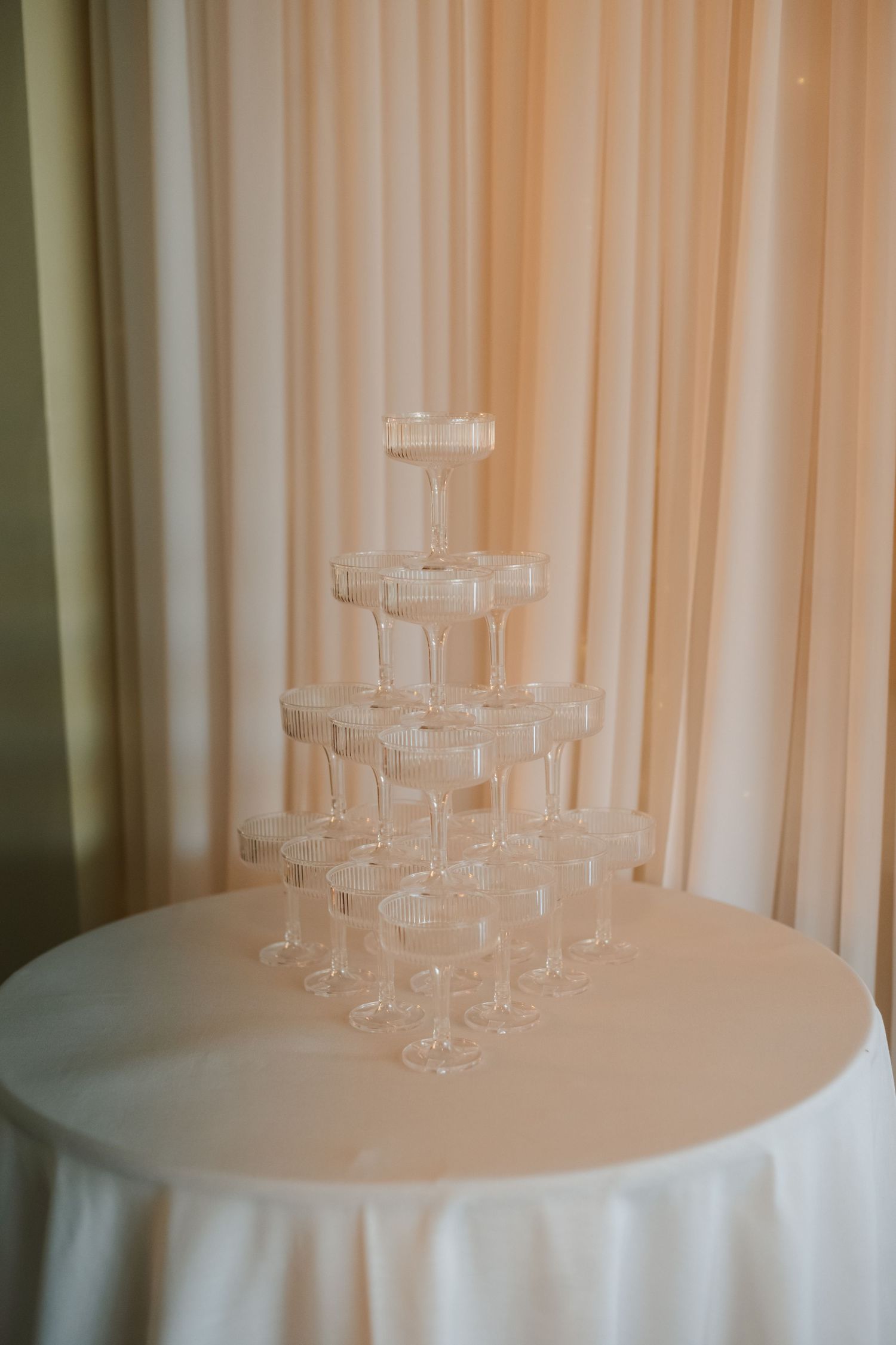 Elegant champagne tower with stacked coupe glasses on a white table against soft pink draping.