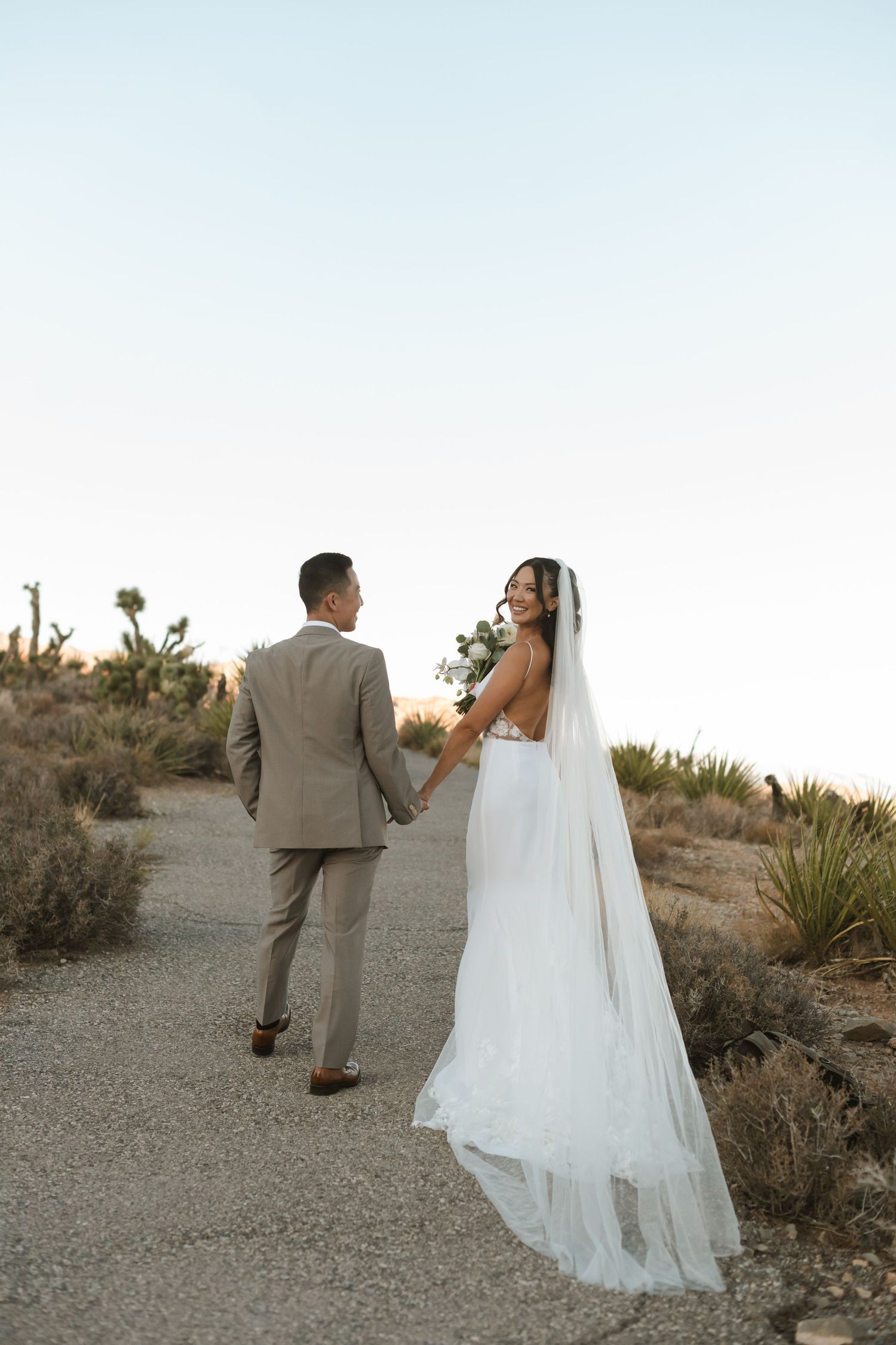 Bride in white gown and veil holds bouquet, walking hand-in-hand with groom in tan suit on desert path.