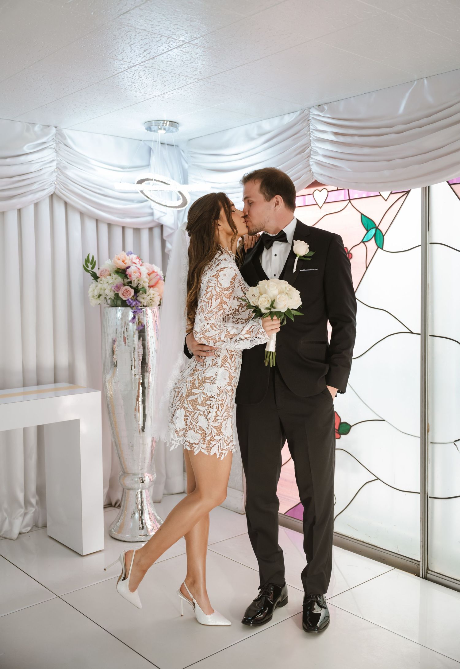 Bride and groom share a kiss in a white chapel, bride wearing lace mini dress holding white bouquet.