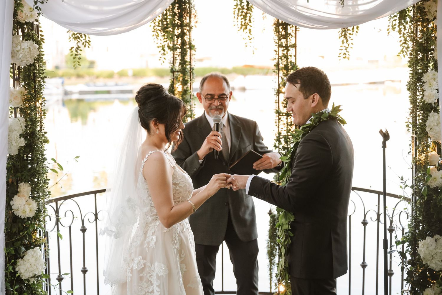 Officiant leads ring exchange between bride and groom at elegant outdoor wedding ceremony with floral arch.