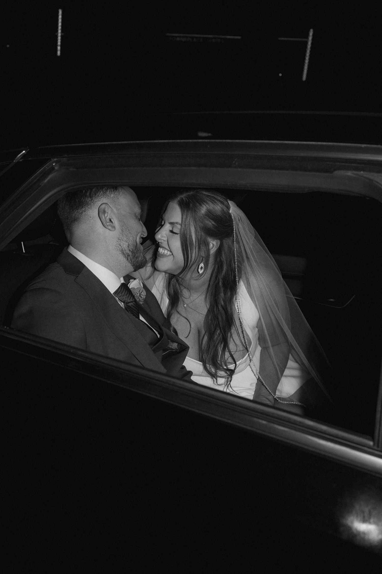 Black and white photo of a smiling bride in a veil and a groom in a suit sharing an intimate moment inside a car at night.