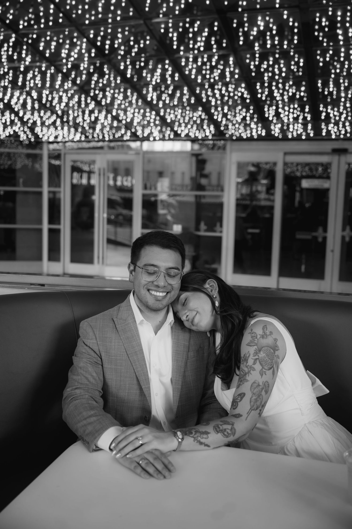 Black and white photo of a smiling couple seated in a booth, her head resting on his shoulder, under a ceiling of twinkling string lights.