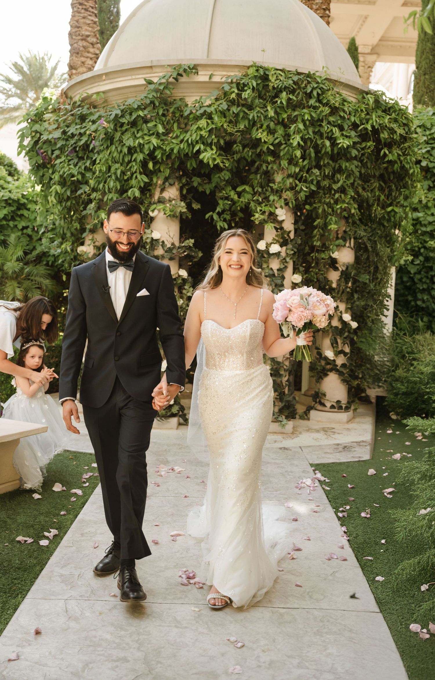 Newlyweds walk hand in hand down a petal-strewn aisle beneath a vine-covered gazebo.