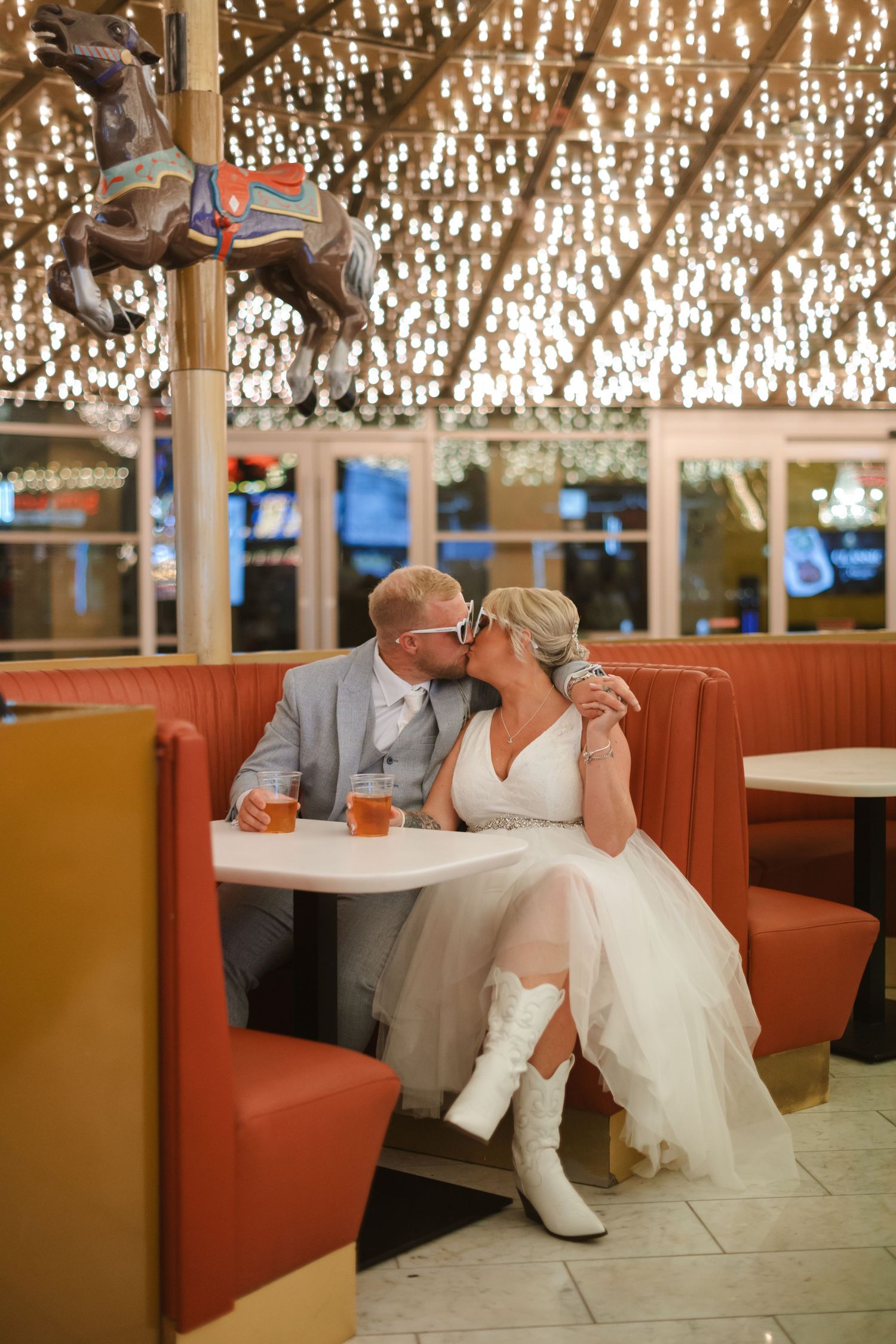 Newlyweds share a kiss in a diner booth, bride in white dress and boots, groom in gray suit, carousel decor above.