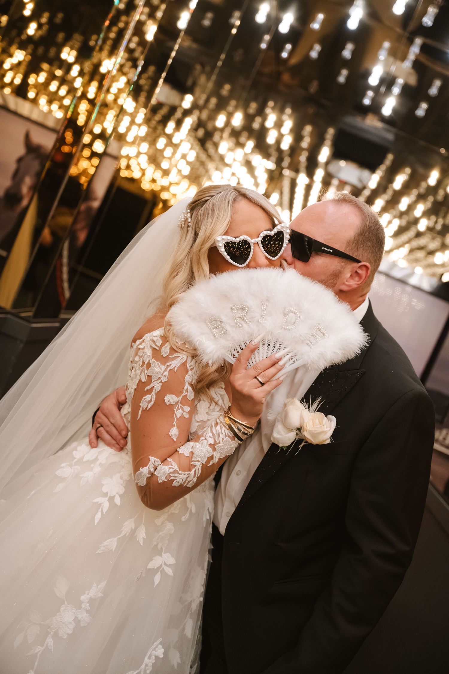 Bride in lace gown and groom in tuxedo kissing behind a white feather fan, wearing heart-shaped sunglasses at wedding reception.