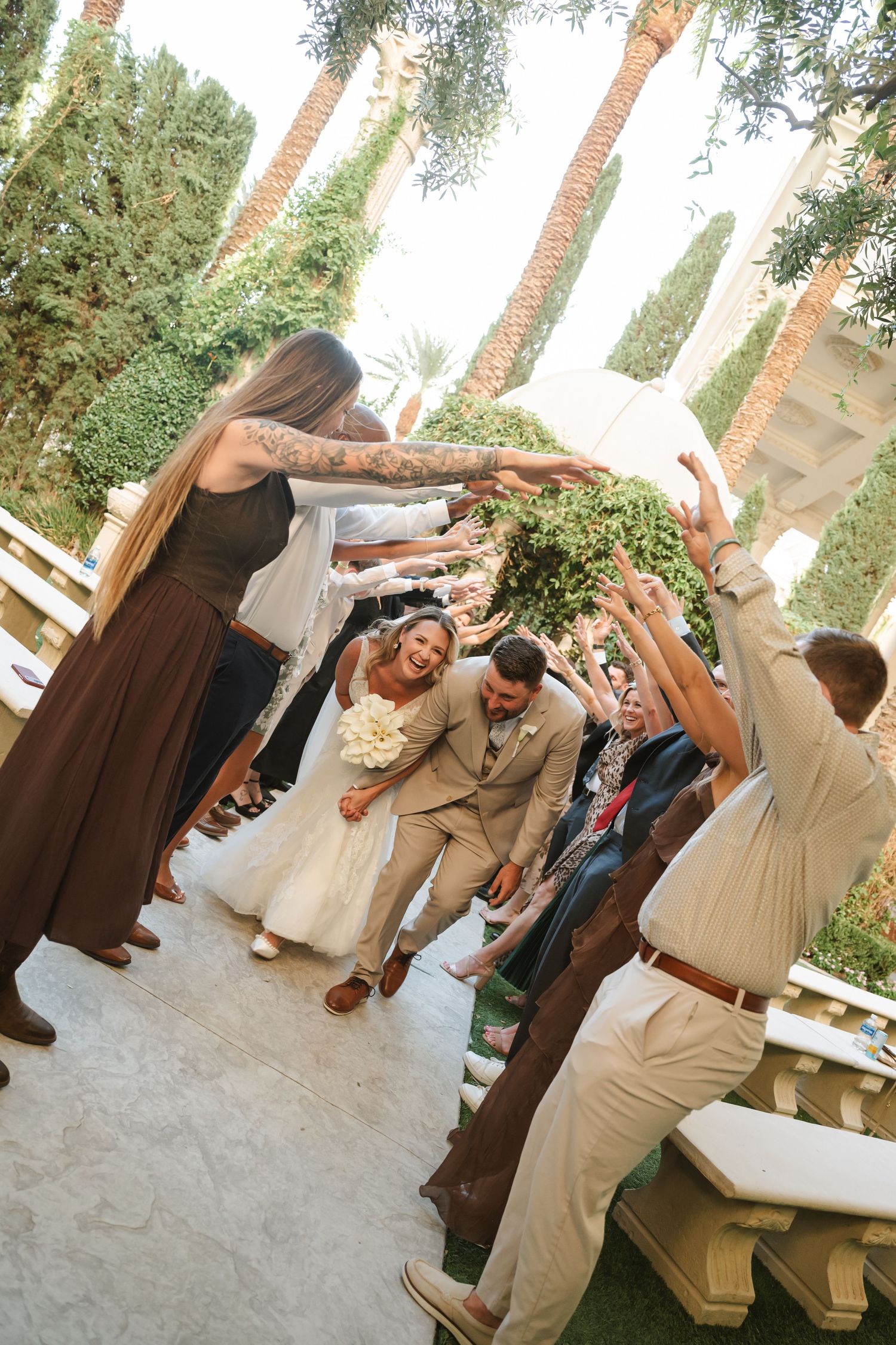 Newlywed couple walks down outdoor wedding aisle under raised arms of guests, bride holds white bouquet.