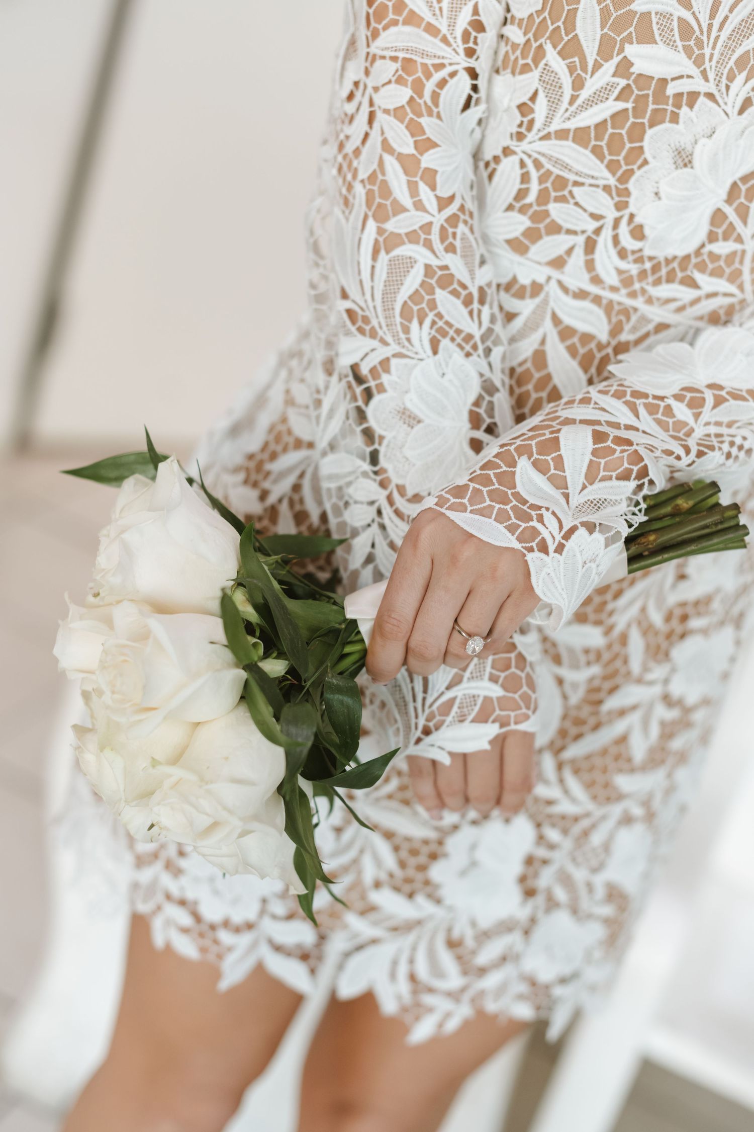 Bride in white lace long-sleeve wedding dress holding elegant white rose bouquet with green leaves.