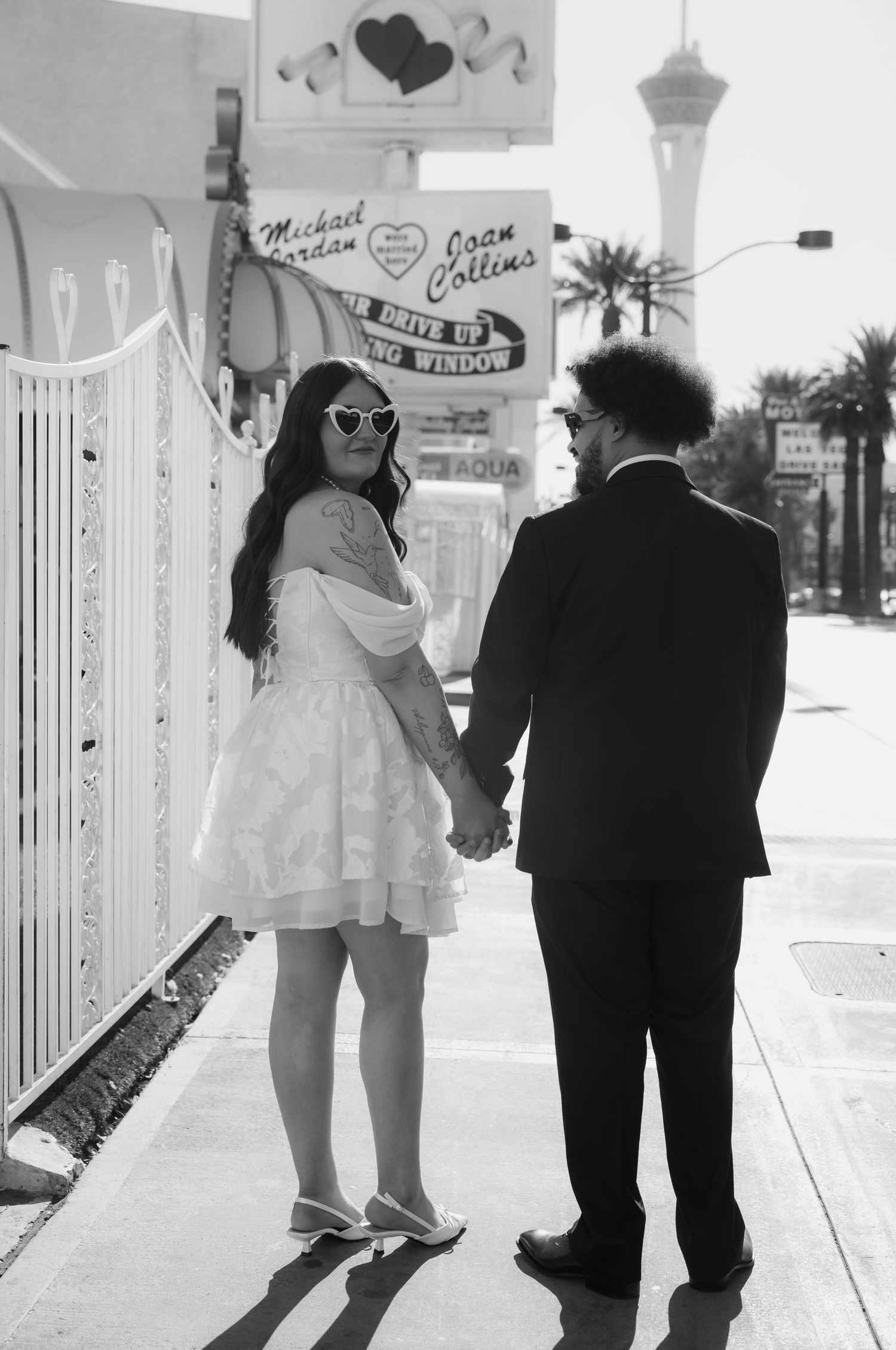 Couple holding hands on Las Vegas Strip sidewalk, woman in white dress with heart sunglasses, man in black suit, black and white photo.