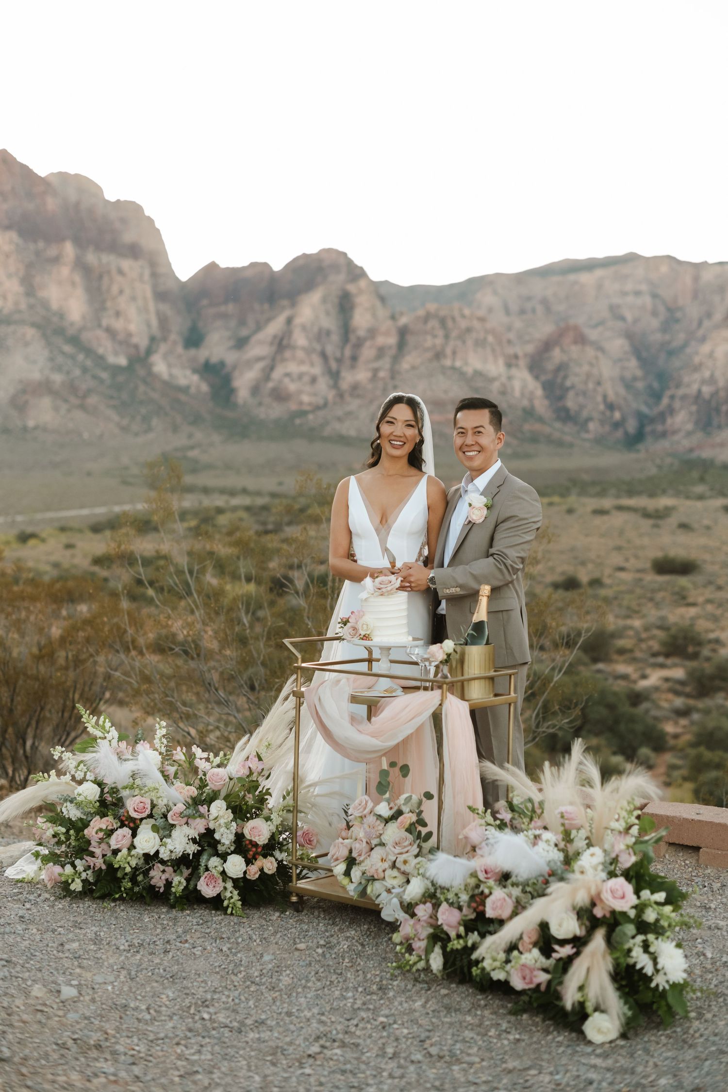 Bride and groom cutting wedding cake outdoors at Red Rock Canyon, surrounded by pink floral arrangements.