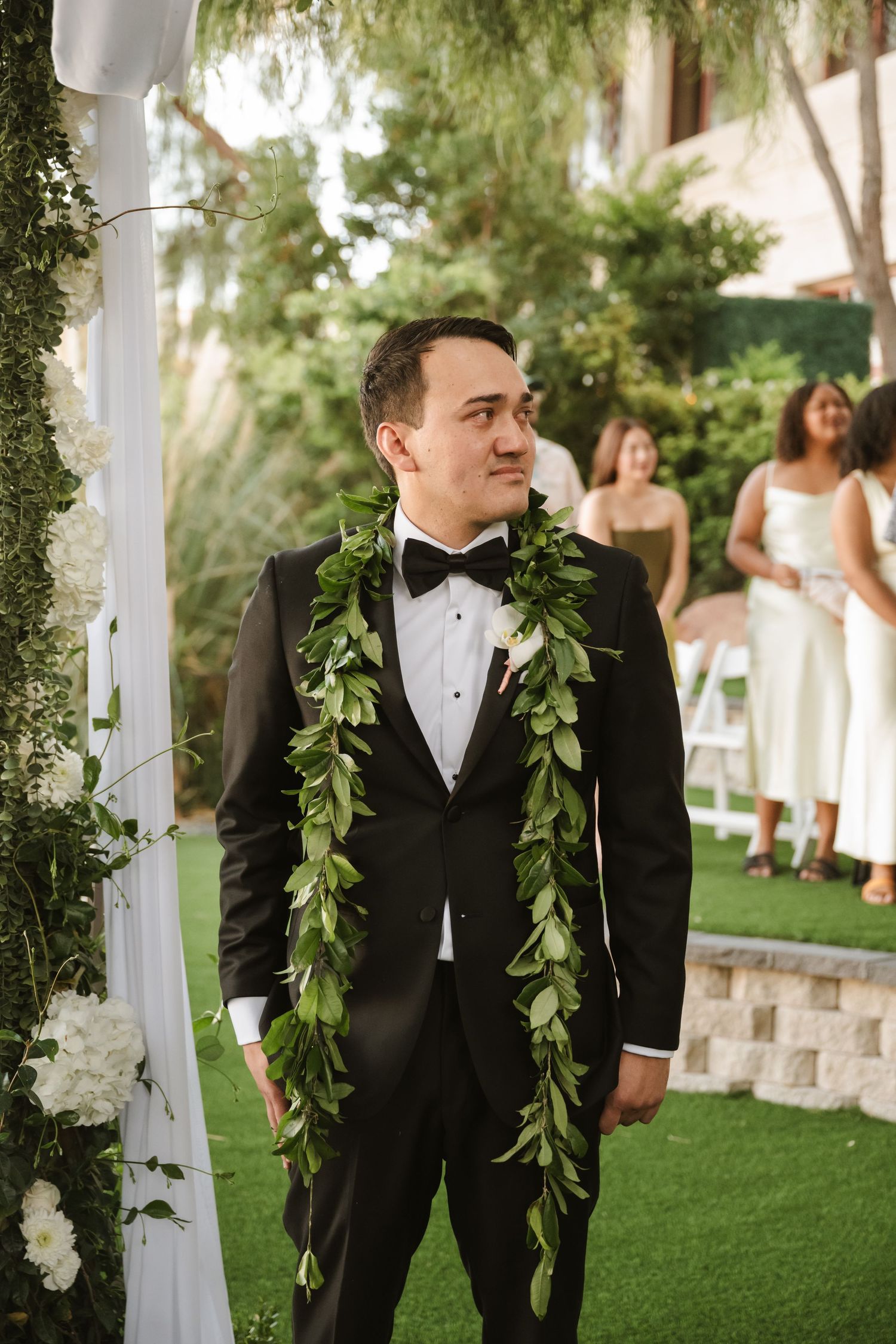 Groom wearing black tuxedo and tropical green lei smiles at outdoor wedding ceremony with guests seated.
