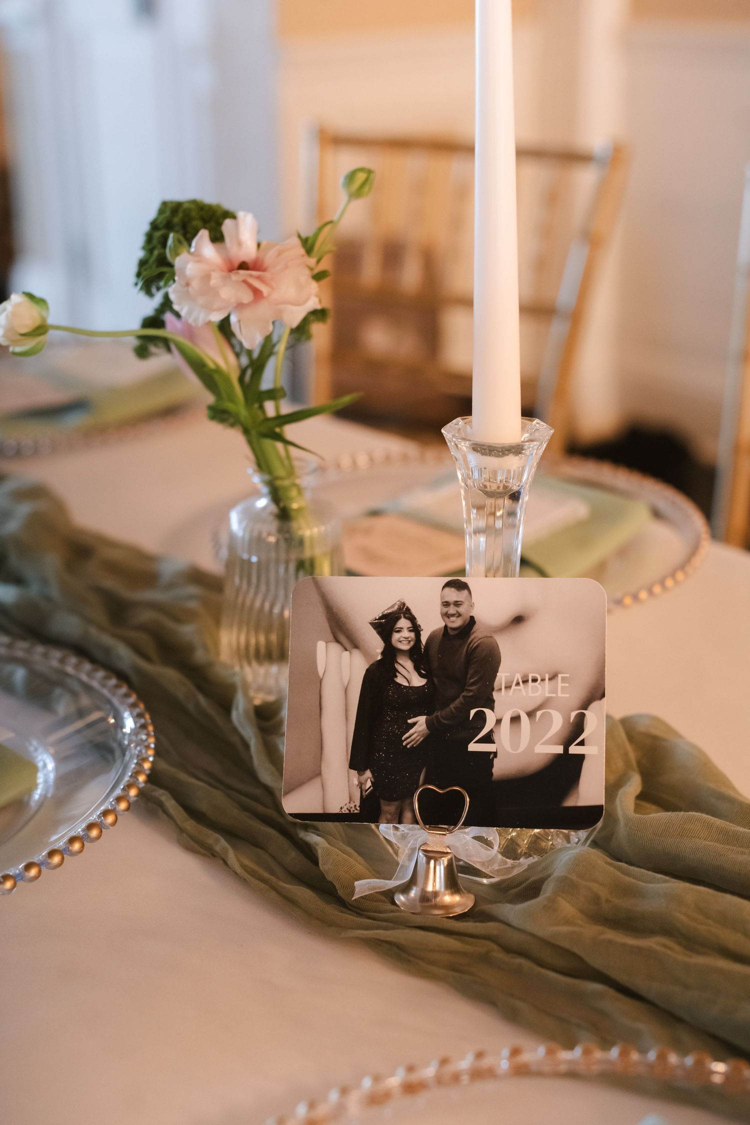 Elegant wedding table setting with a 2022 photo card, crystal candlestick, pink flowers, and sage green cheesecloth runner.